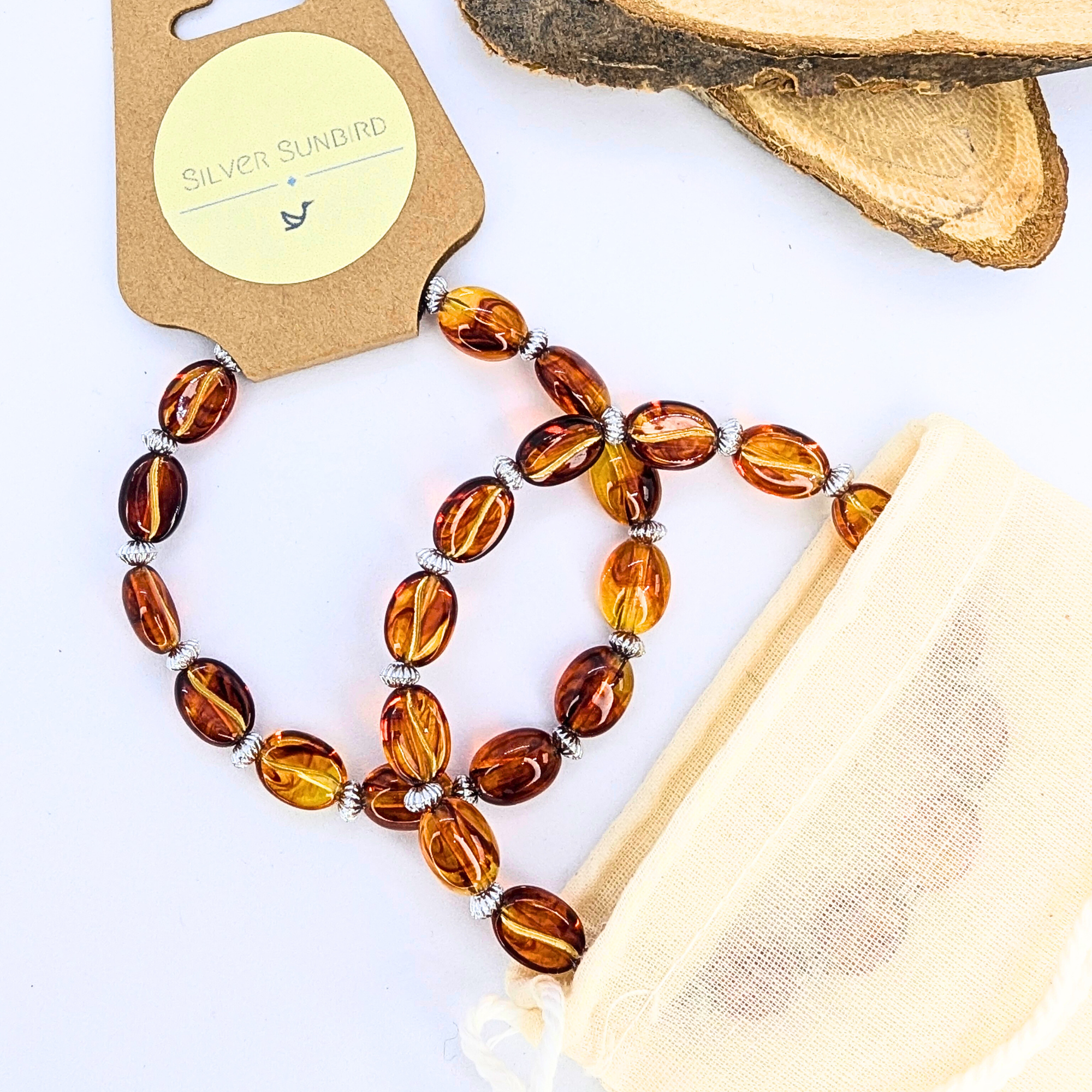 Displayed on a white surface, an amber beaded necklace with silver spacers lies beside a beige "Silver Sunbird" pouch. Wood slices in the background add charm reminiscent of Silver Sunbird&