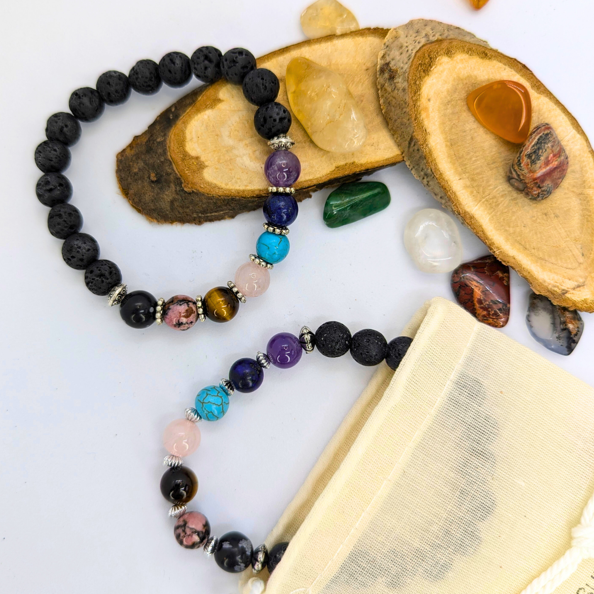 The Chakra Bracelet by Silver Sunbird features black and colorful semi-precious gemstones, displayed on wood slices with tumbled crystals and a beige pouch, all set on a white surface.