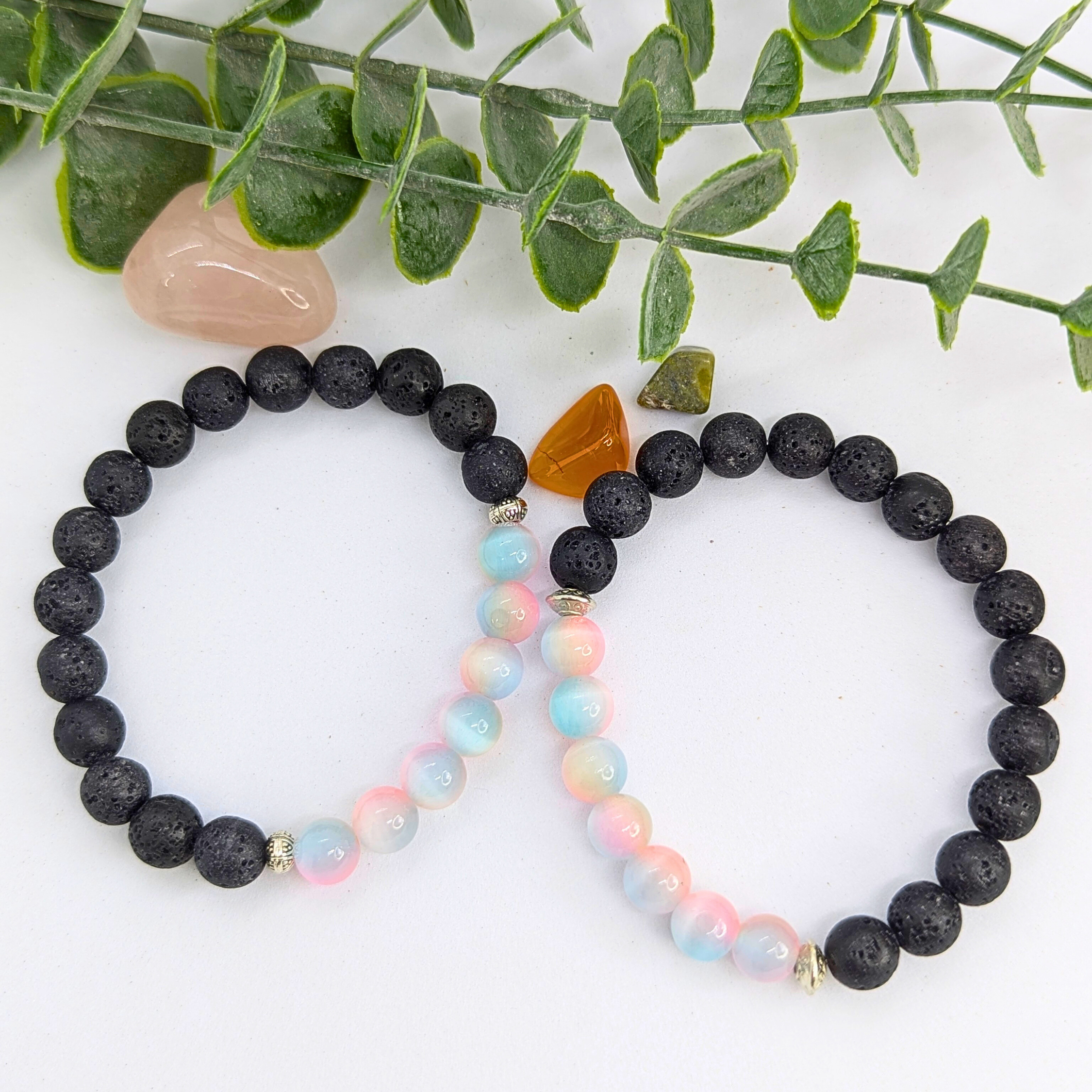 A pastel beaded bracelet and the Silver Sunbird Mermaid-Inspired Lava Stone Bracelet with black beads and a brownish-orange stone rest on a white surface, with green leaves and polished stones in the background.