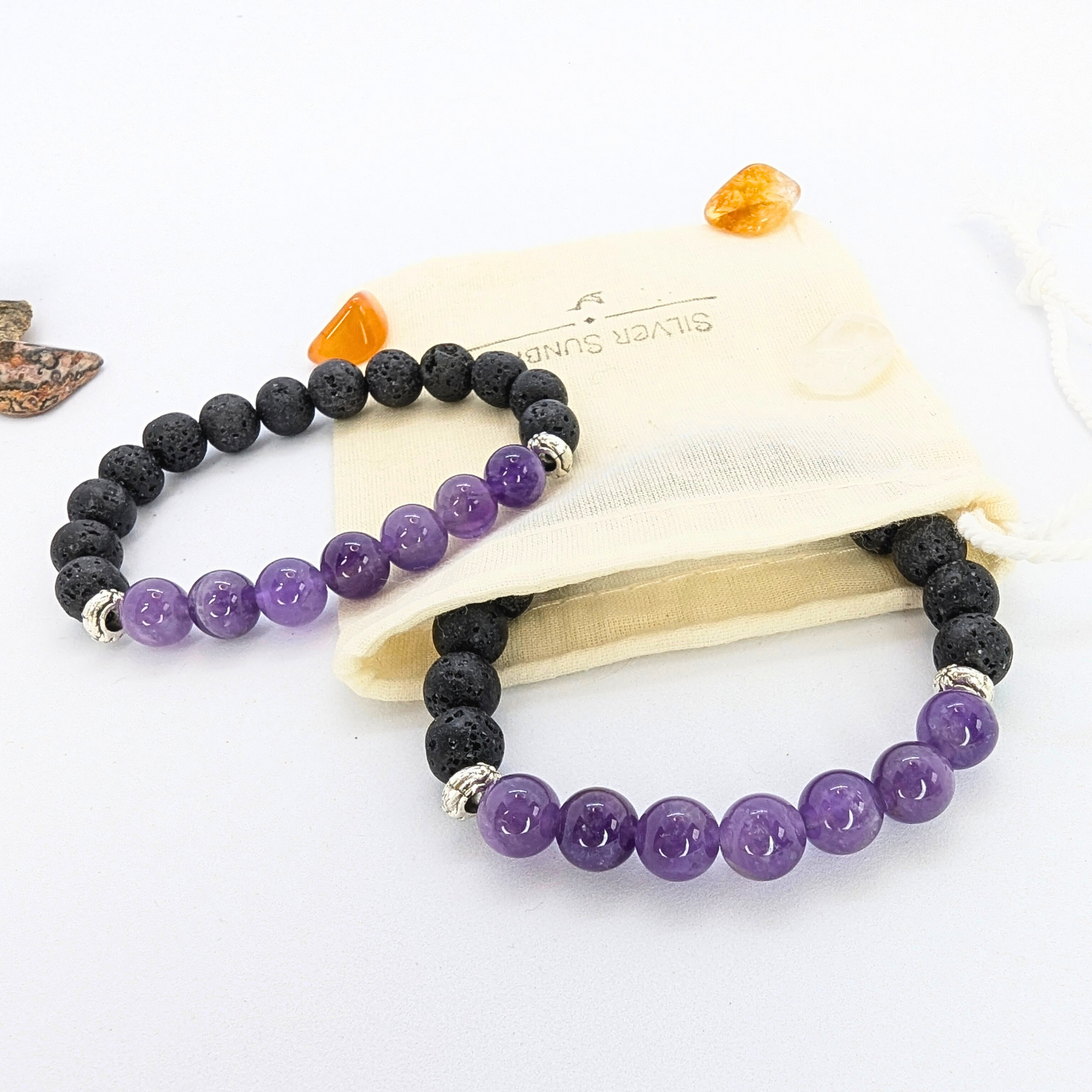 Two stretch bracelets featuring black lava stones and purple amethyst beads are displayed on a wooden slice. A beige drawstring pouch with "Silver Surfer" branding is positioned to the side, along with small polished stones in orange and brown hues. The amethyst beads are glossy and smooth, contrasting with the porous texture of the lava stones.