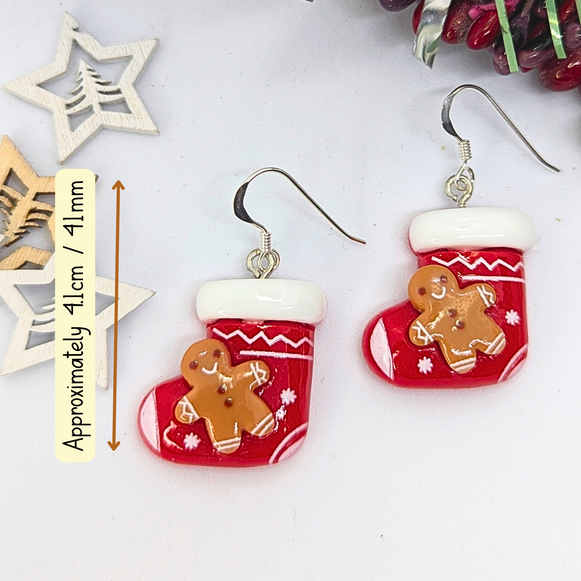 The Gingerbread Men Stocking Earrings by Silver Sunbird are designed in the shape of red Christmas stockings with gingerbread men patterns. They feature silver hooks and measure approximately 4.1 cm long. Festive wooden star decorations provide a charming backdrop for these unique earrings.