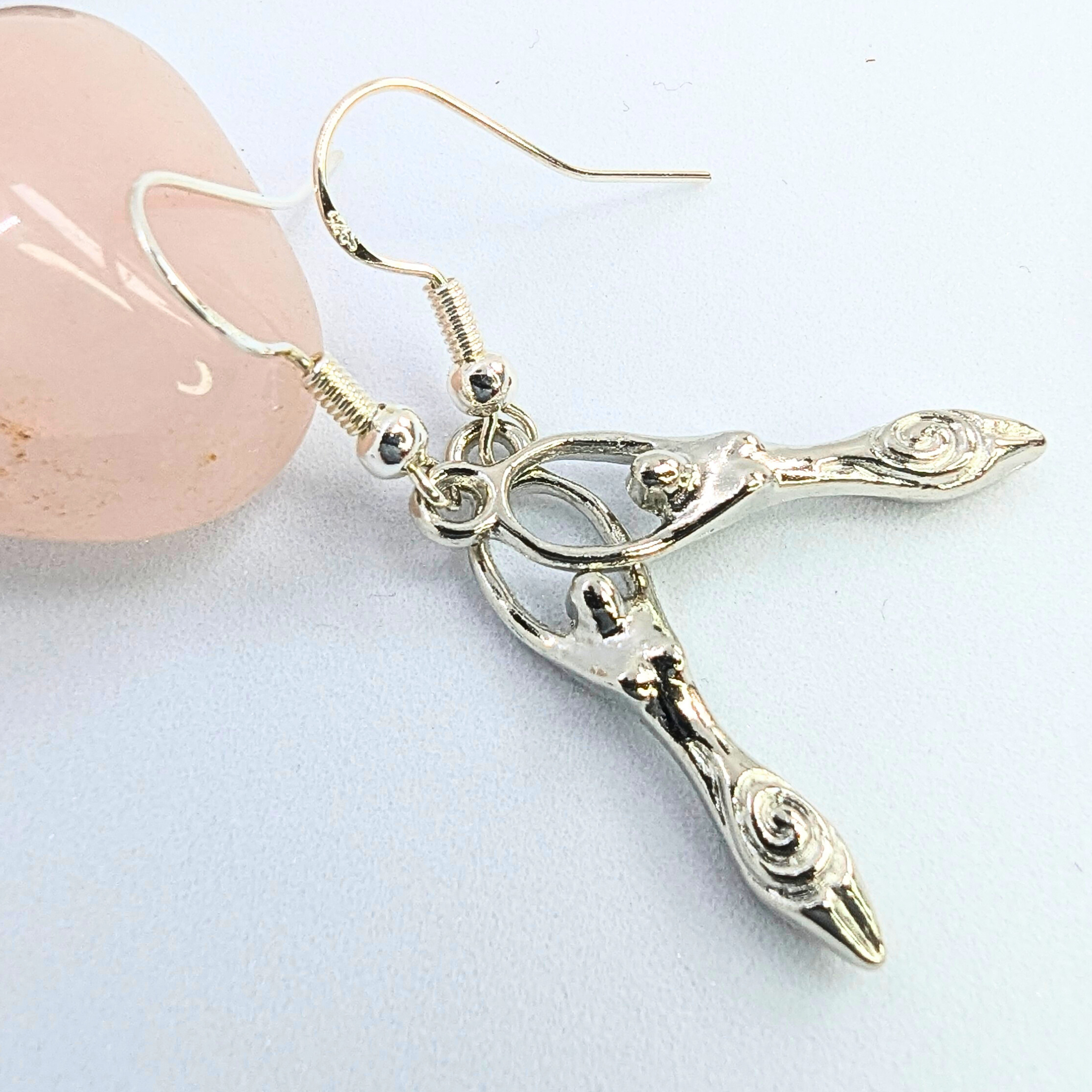 The Silver Sunbird Fertility Goddess Dangle Earrings, featuring spiral accents in sterling silver, are displayed beside a pink polished stone on a white background, showcasing this symbolic piece of jewelry.