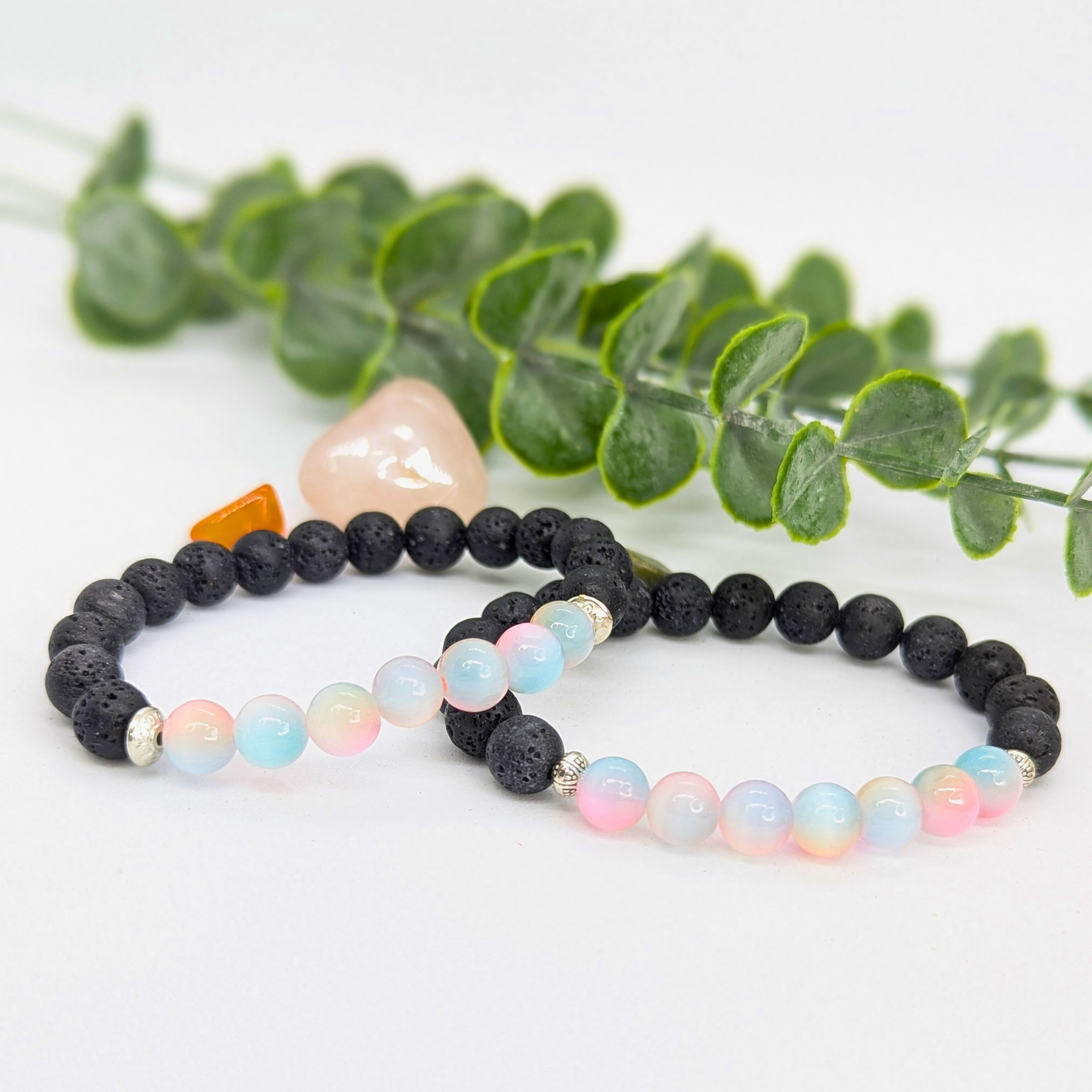Silver Sunbird’s Mermaid-Inspired Lava Stone Bracelet is displayed on a white surface beside a green leafy branch and two polished stones—an ideal accessory for ocean lovers.