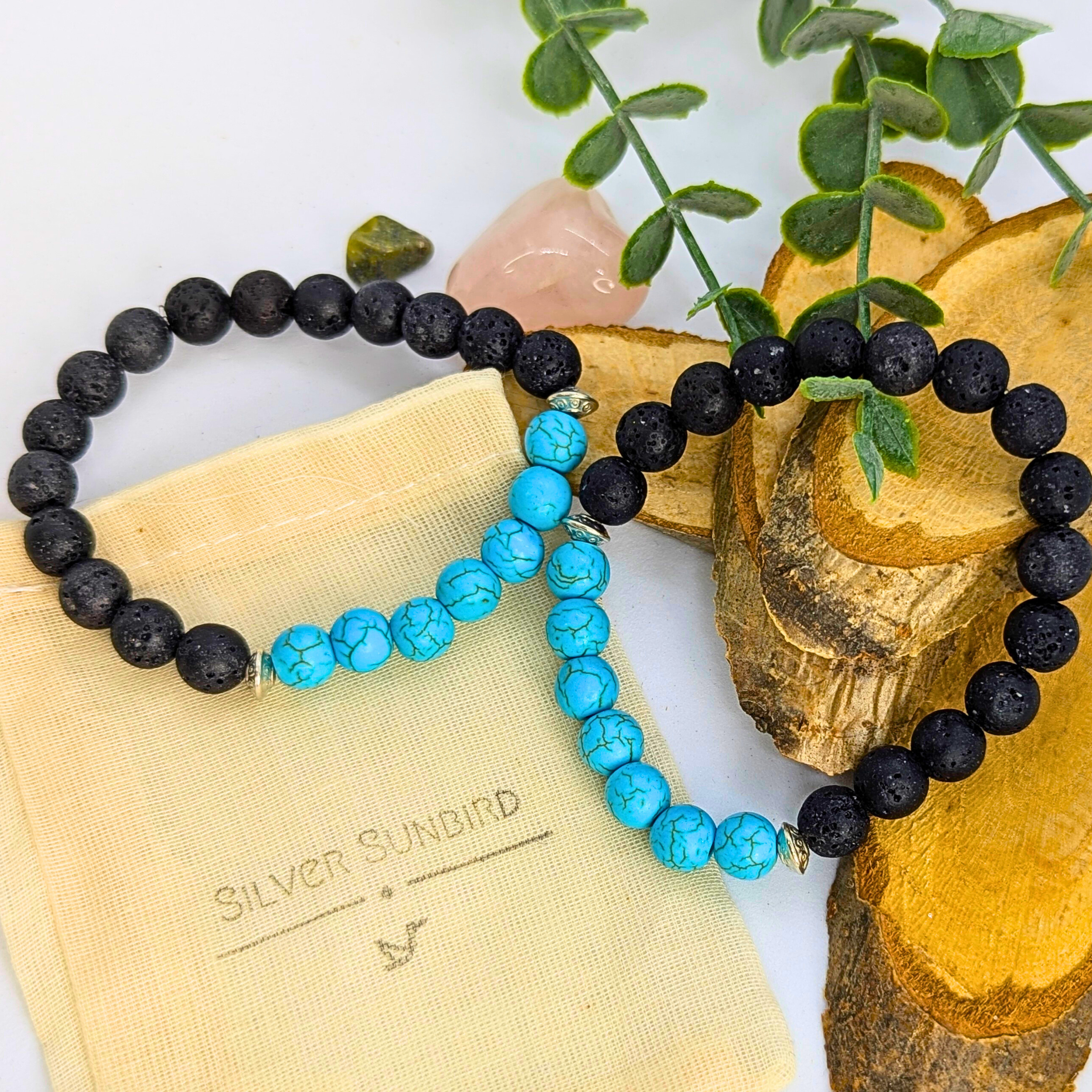 Two Turquoise and Lava Stone Bracelets by Silver Sunbird—one all-black, one with alternating turquoise and black beads—are displayed on a cream pouch labeled "Silver Sunbird," with green leaves and wooden slices in the background.