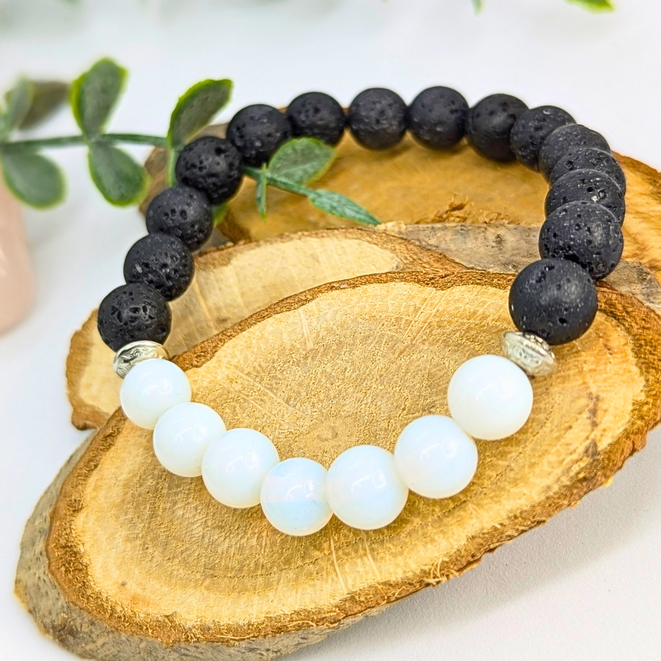 The Silver Sunbird Opal and Lava Stone Bracelet features alternating black lava stones and smooth white opal beads, showcased on a wooden slice with green leaves in the background.