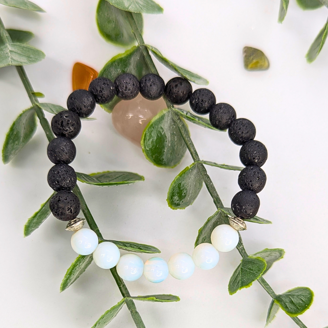 The Silver Sunbird Opal and Lava Stone Bracelets are displayed on green leafy stems, surrounded by small polished stones, against a white background.