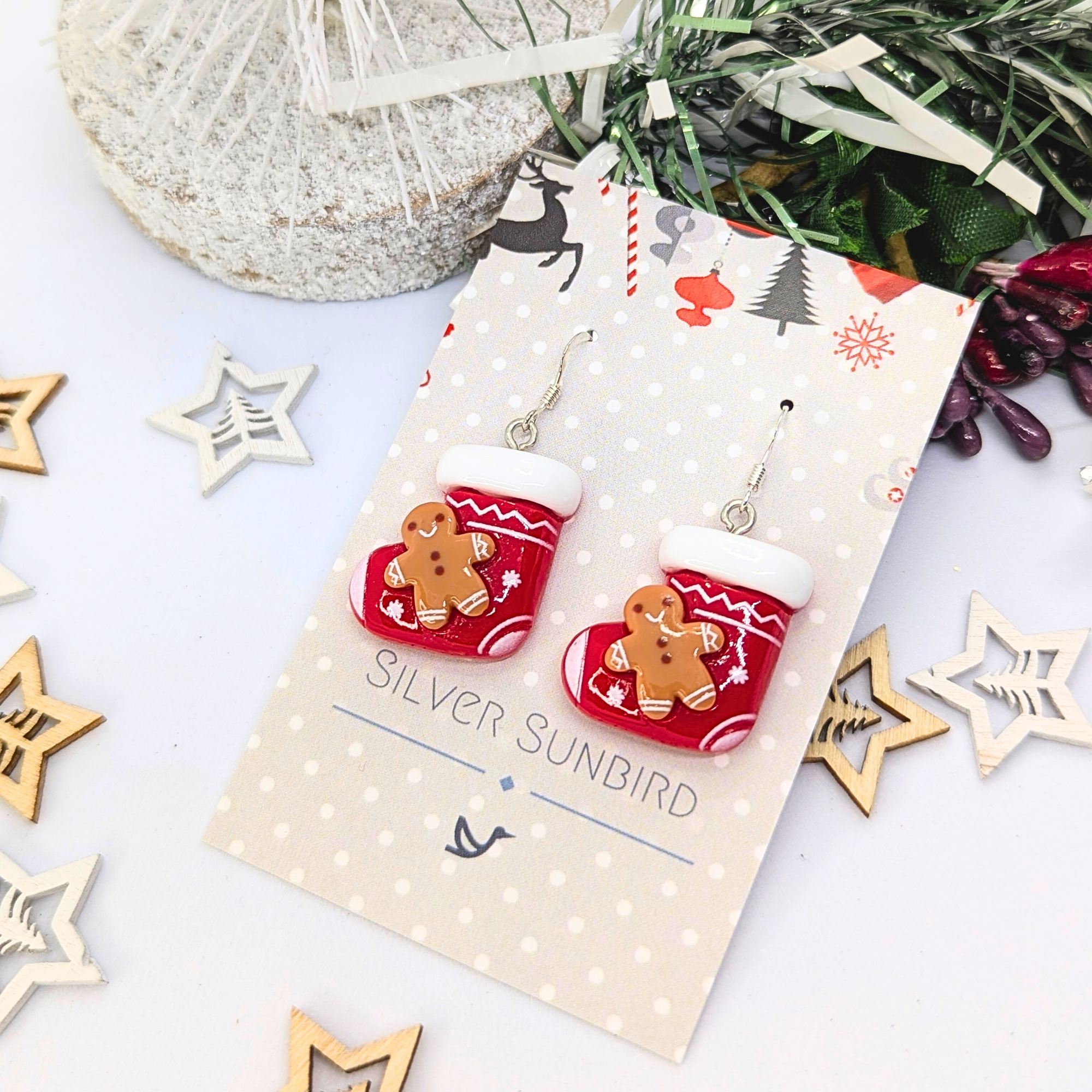 Gingerbread Men Stocking Earrings by Silver Sunbird are showcased on a cardboard backing, accompanied by wooden star ornaments and a snowy decorative branch.