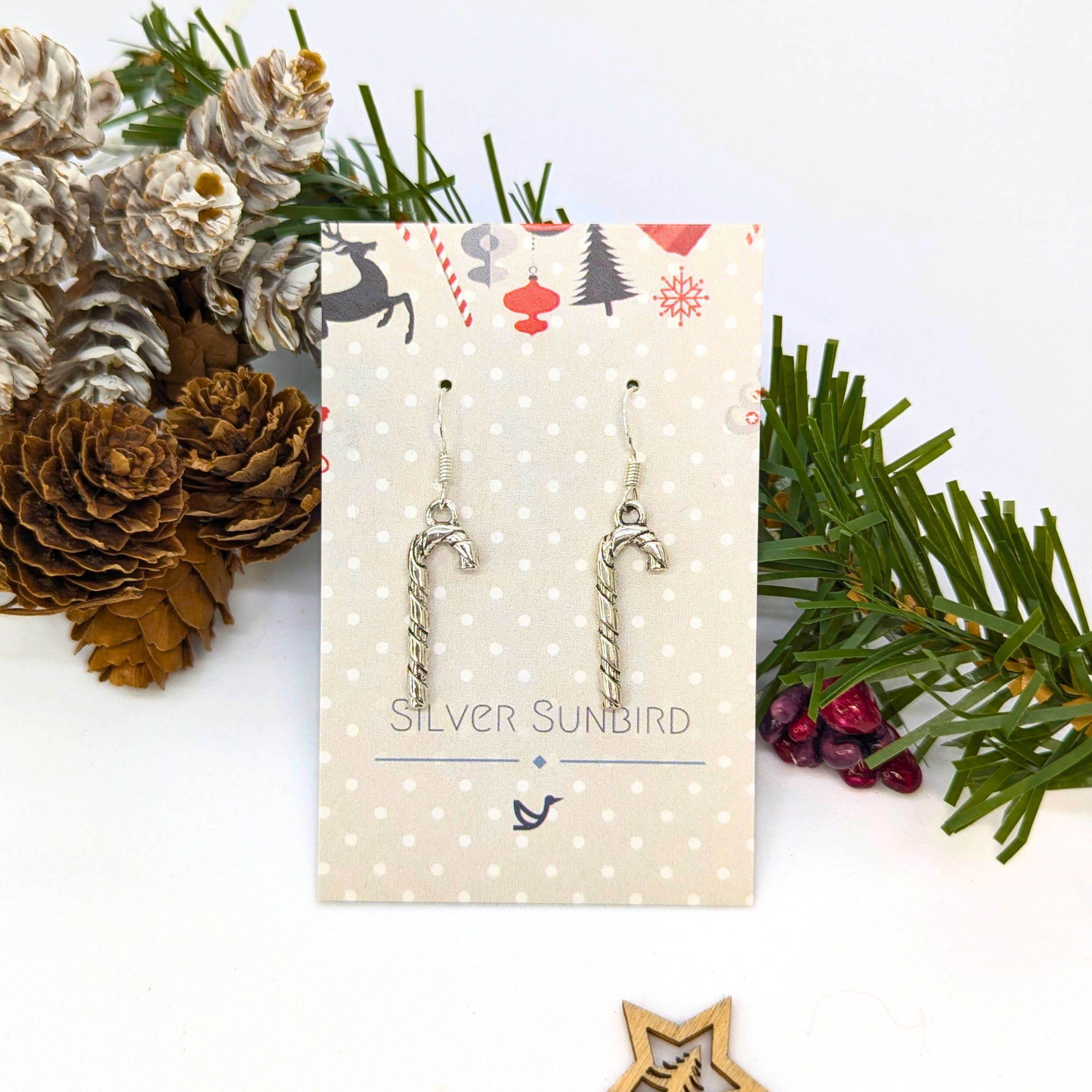 Classic Candy Cane Earrings by Silver Sunbird hang from a branded card, surrounded by pinecones, evergreen branches, and festive holiday décor—perfect Christmas jewelry for the season.