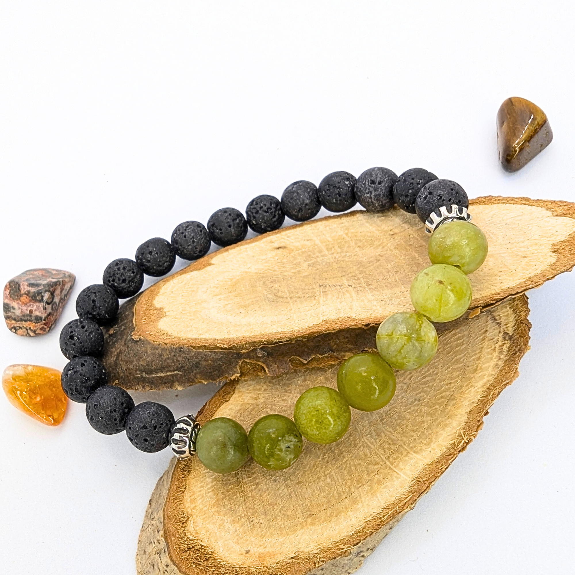 The Silver Sunbird Peridot and Lava Stone Bracelets, featuring alternating green and black stone beads with decorative metal accents, are displayed on stacked wood beside polished amber, brown, and gray stones against a white background.