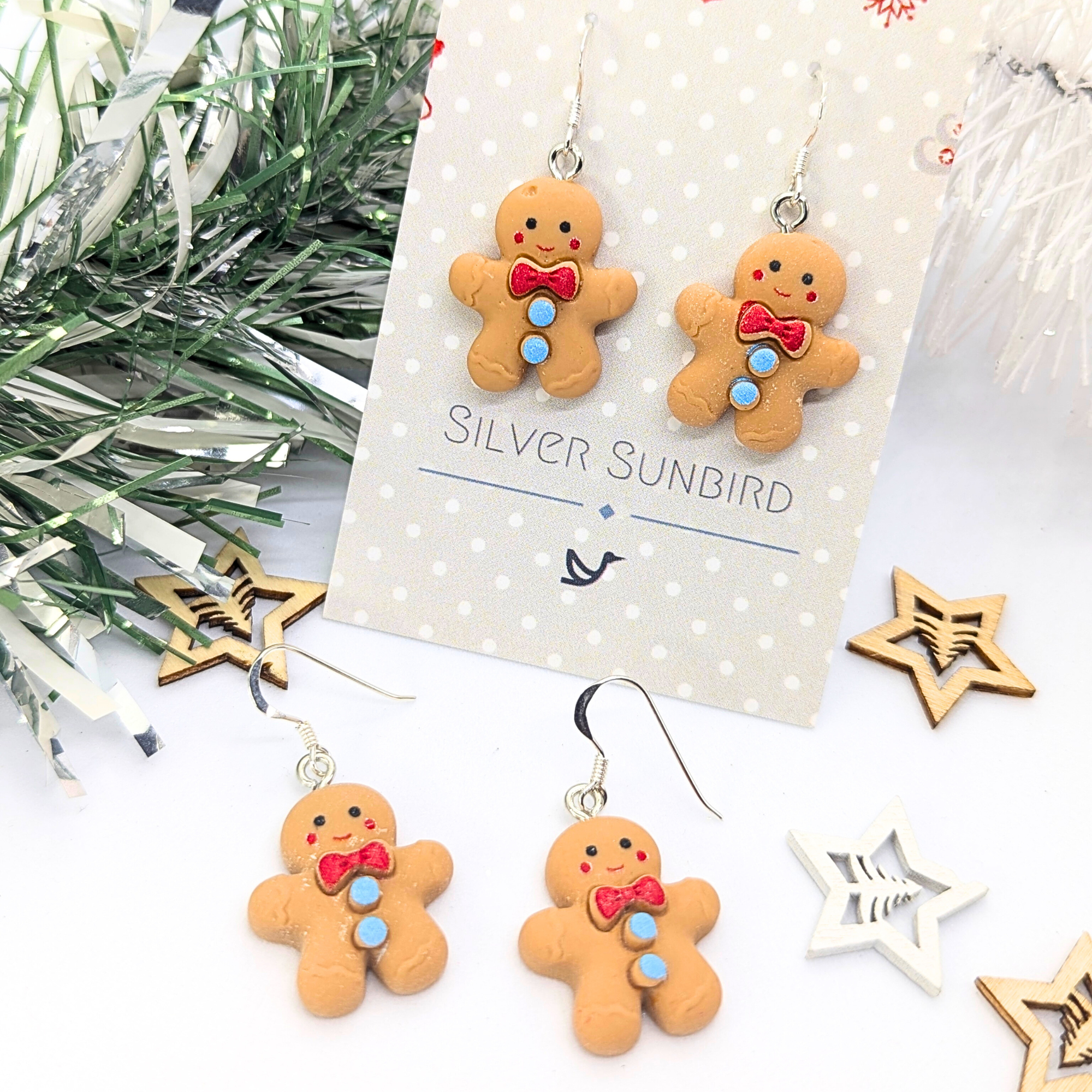 Cheerful Gingerbread Man Earrings by Silver Sunbird, featuring charming gingerbread men adorned with red bows and blue buttons, are presented on a card surrounded by decorative stars and festive greenery.
