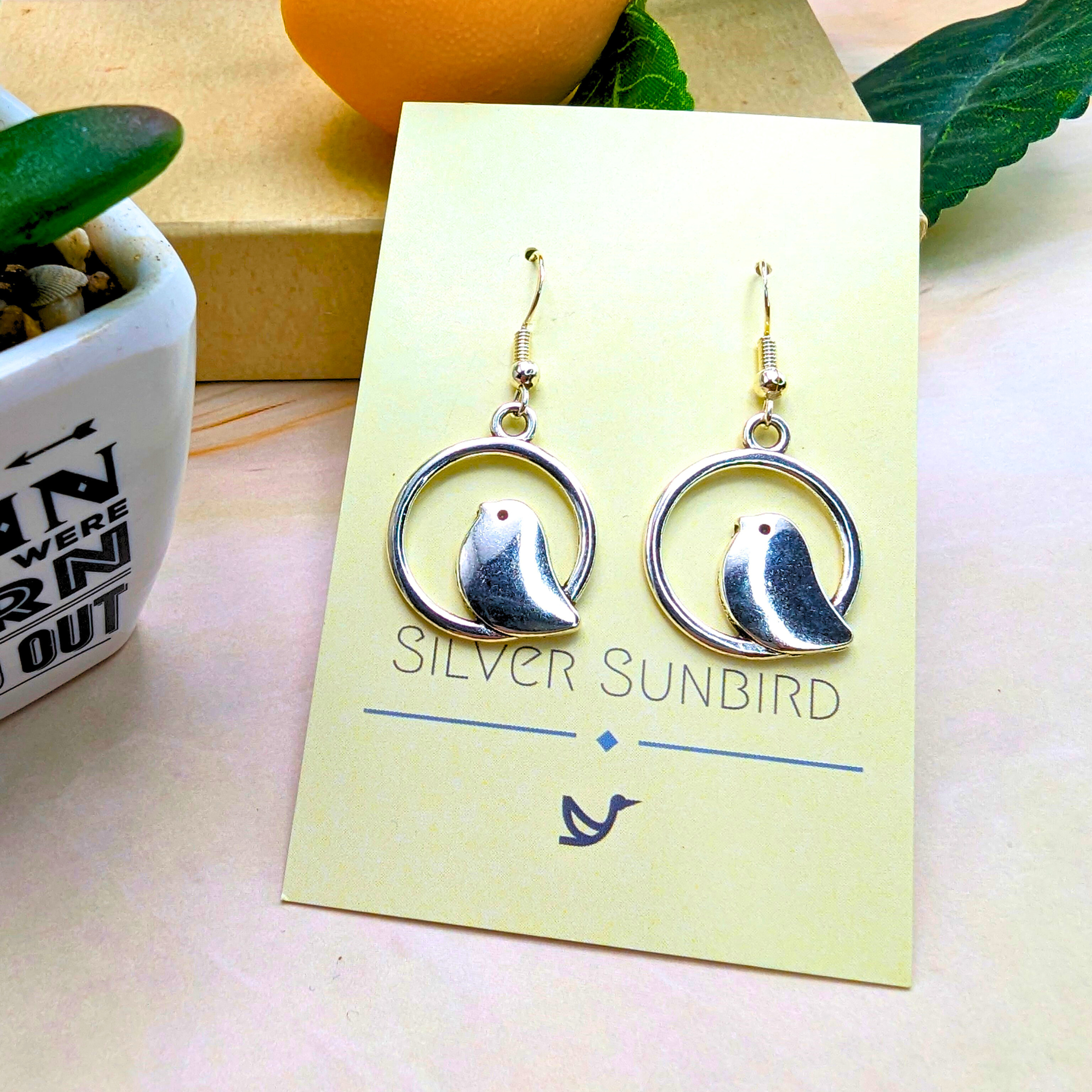 Displayed on a Silver Sunbird card, the Beautiful Bird Earrings are crafted from 925 Sterling Silver, showcasing a delicate bird in circular frames. A small plant and decor enhance the elegant presentation, perfect for bird lovers.