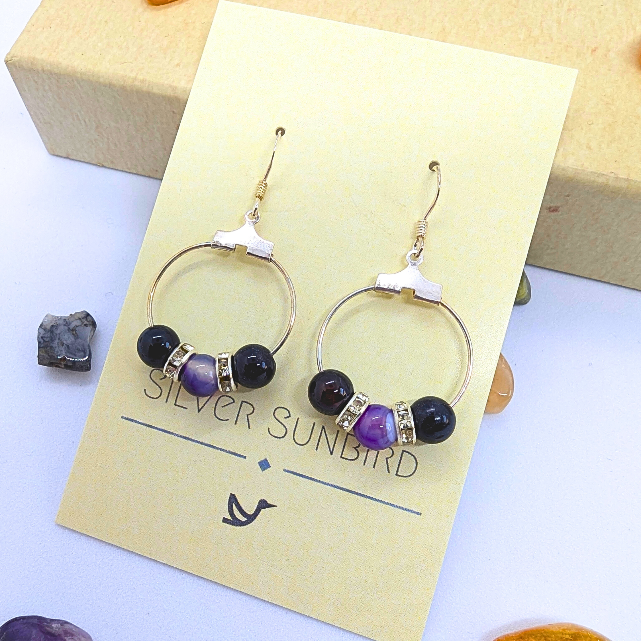 A pair of circular silver earrings embellished with purple gemstones, featuring Amethyst dangle elements and silver rhinestones, are displayed on a yellow card labelled "Silver Sunbird." The card is placed on a light surface next to scattered decorative stones.
