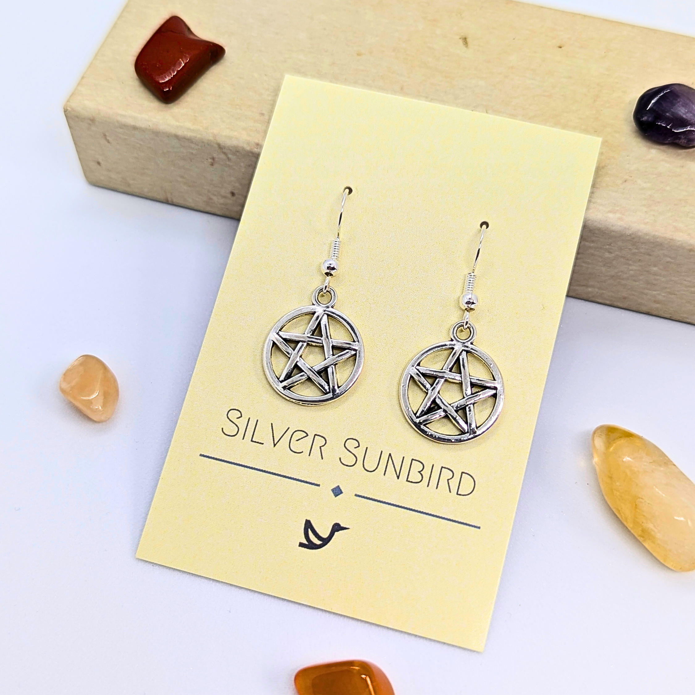 Pentagram Earrings by Silver Sunbird dangle elegantly on a yellow card, surrounded by colorful stones and a box—ideal Wicca jewelry for those who love pagan symbolism.