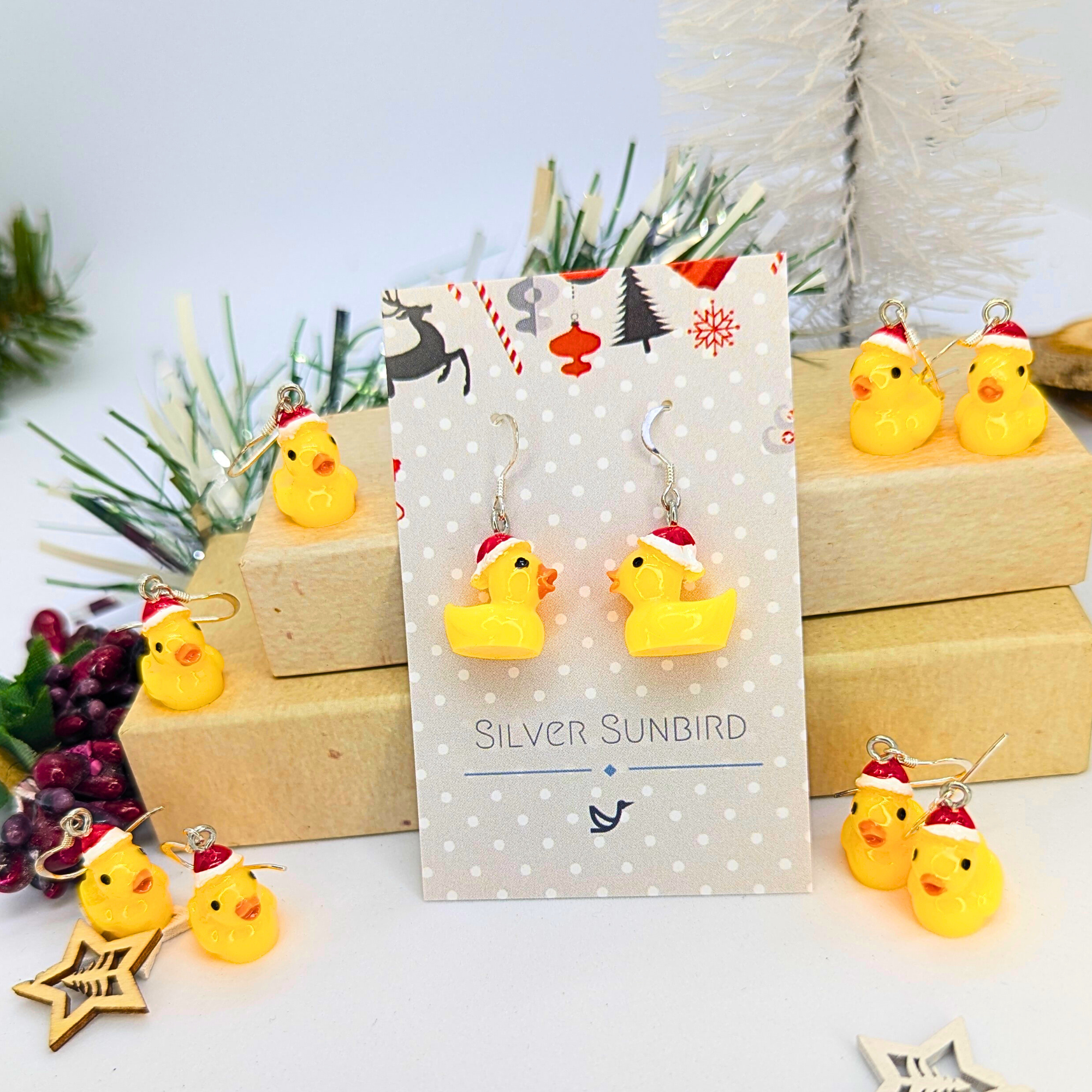 A festive arrangement featuring Festive Duck Earrings from Silver Sunbird, crafted in sterling silver and decorated with Santa hats, presented on a polka-dot card. The display is enhanced by additional duck decorations and holiday-themed items placed on wooden boxes.