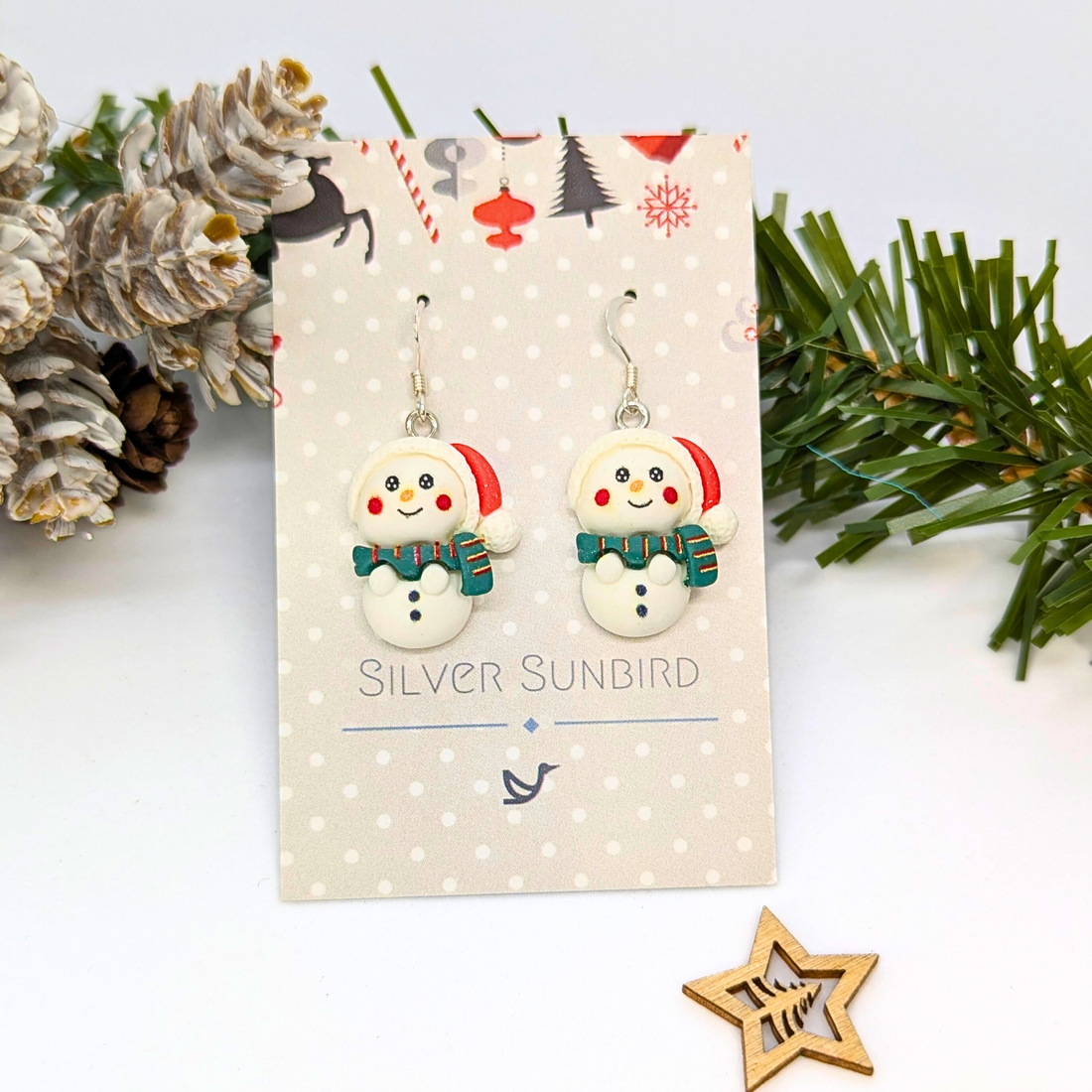 The Bright Snowmen Earrings by Silver Sunbird feature red hats and green scarves, displayed on a festive card with pine, pinecones, and a wooden star. These cheerful Christmas earrings add holiday flair. Card reads "SILVER SUNBIRD.