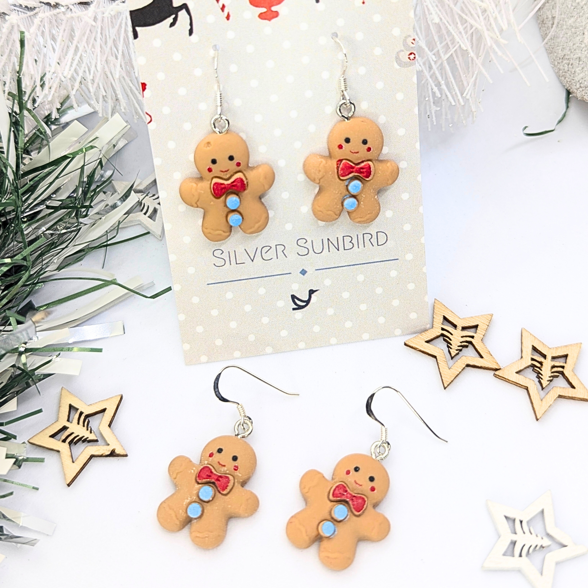 Displayed on a white card labeled "Silver Sunbird," the Cheerful Gingerbread Man Earrings are adorned with red bow ties and blue buttons. The background features festive decor, including white branches and wooden star ornaments.