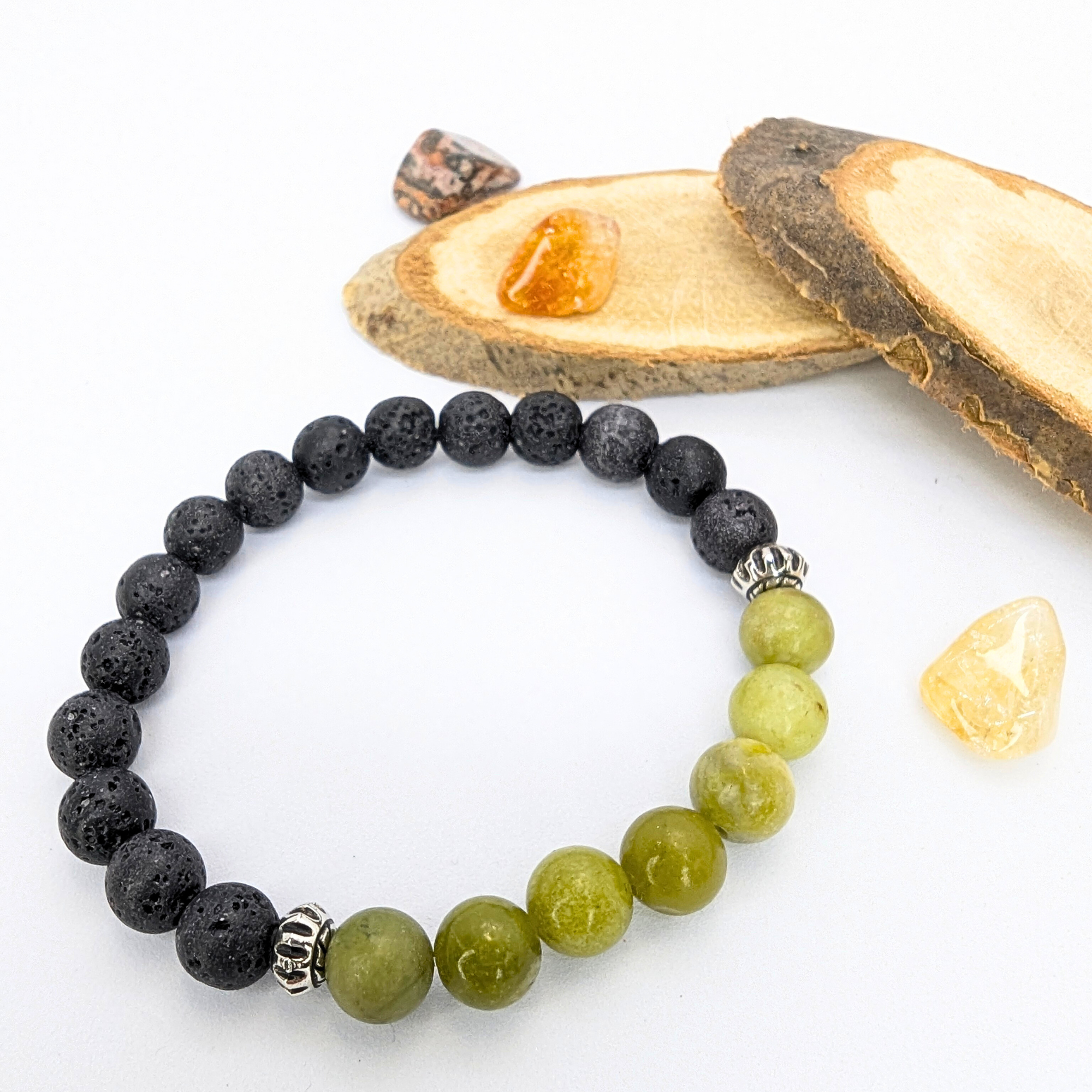 The Silver Sunbird Peridot and Lava Stone Bracelets feature black lava and green peridot beads with silver spacers, displayed on a white surface next to polished stones and wooden slices.