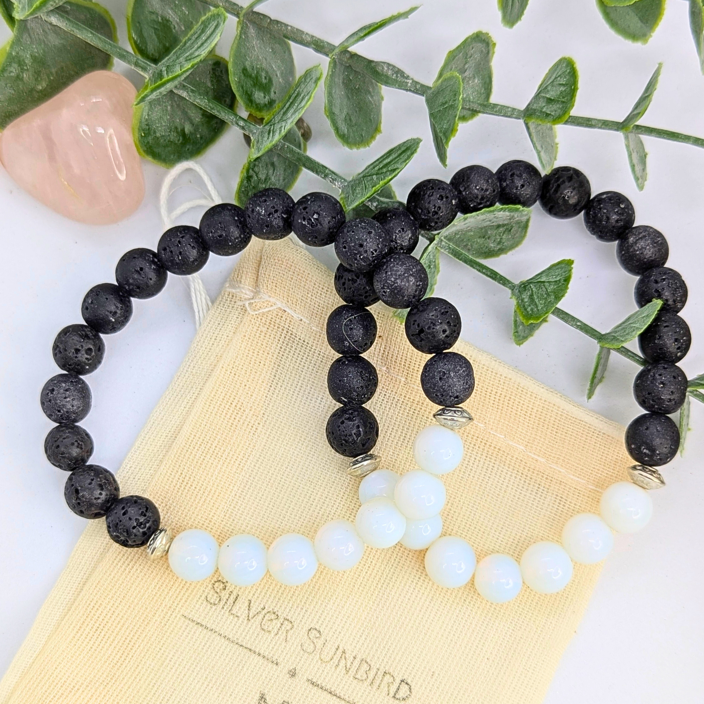 The Opal and Lava Stone Bracelets by Silver Sunbird rest on a beige pouch with green leaves and a pink stone in the background.