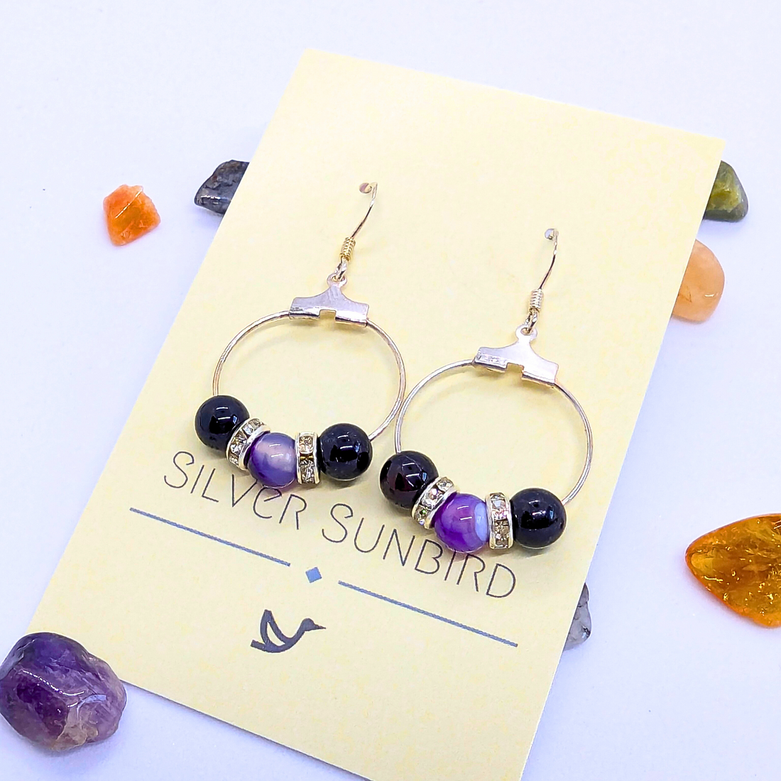 A pair of Amethyst Statement Large Earrings adorned with purple gemstones and silver rhinestone spacers is displayed on a card labelled "Silver Sunbird." The card is set against a white background, scattered with small natural gemstones in orange, green, and purple hues.