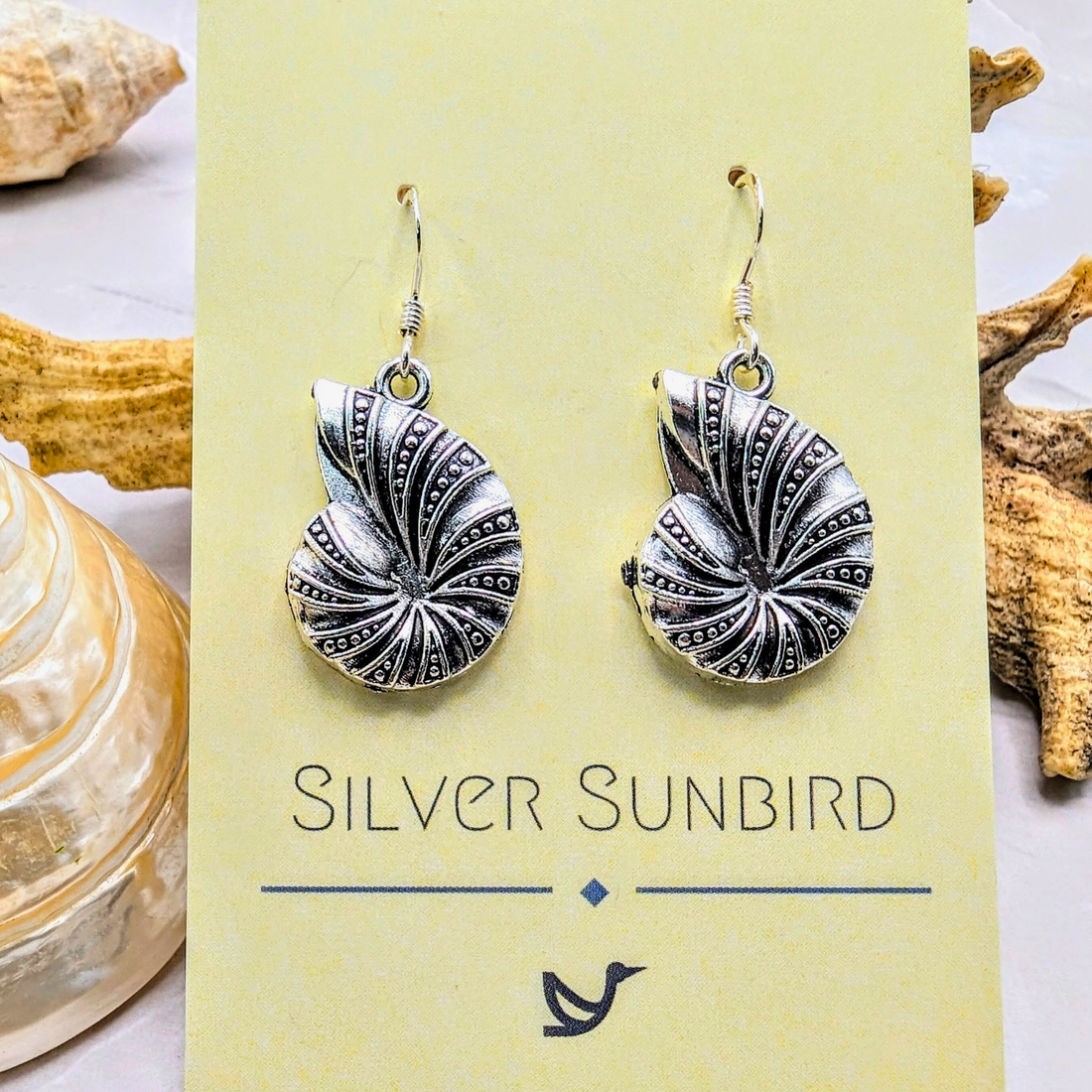 A pair of Shoreline Spiral Shell Earrings by Silver Sunbird—sterling silver, nautilus shell-shaped—displayed on a branded card, with seashells and driftwood in the background.