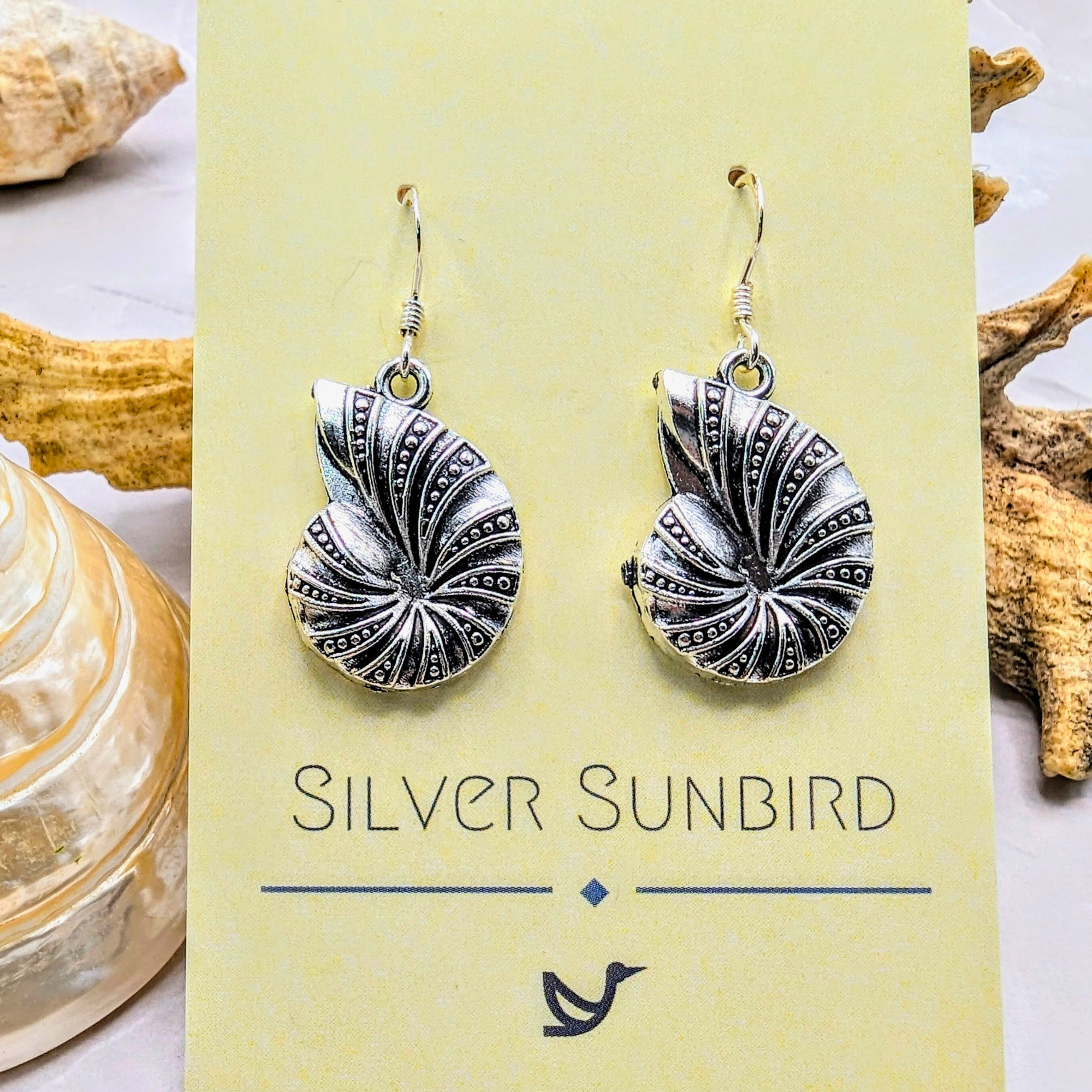 A pair of Shoreline Spiral Shell Earrings by Silver Sunbird—sterling silver, nautilus shell-shaped—displayed on a branded card, with seashells and driftwood in the background.