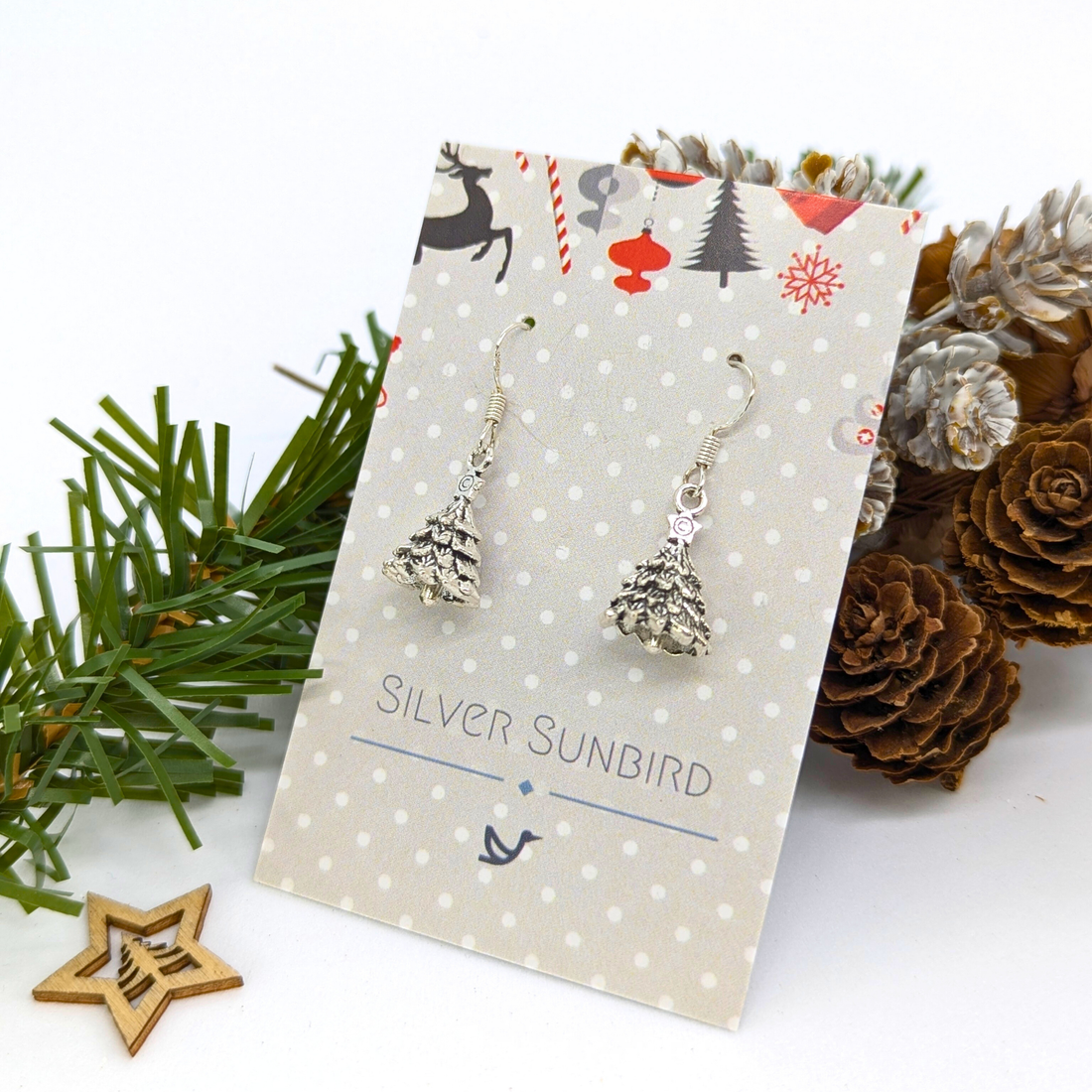 A pair of 3D Christmas Tree Earrings by Silver Sunbird, displayed on a branded card with pinecones, greenery, and a wooden star.