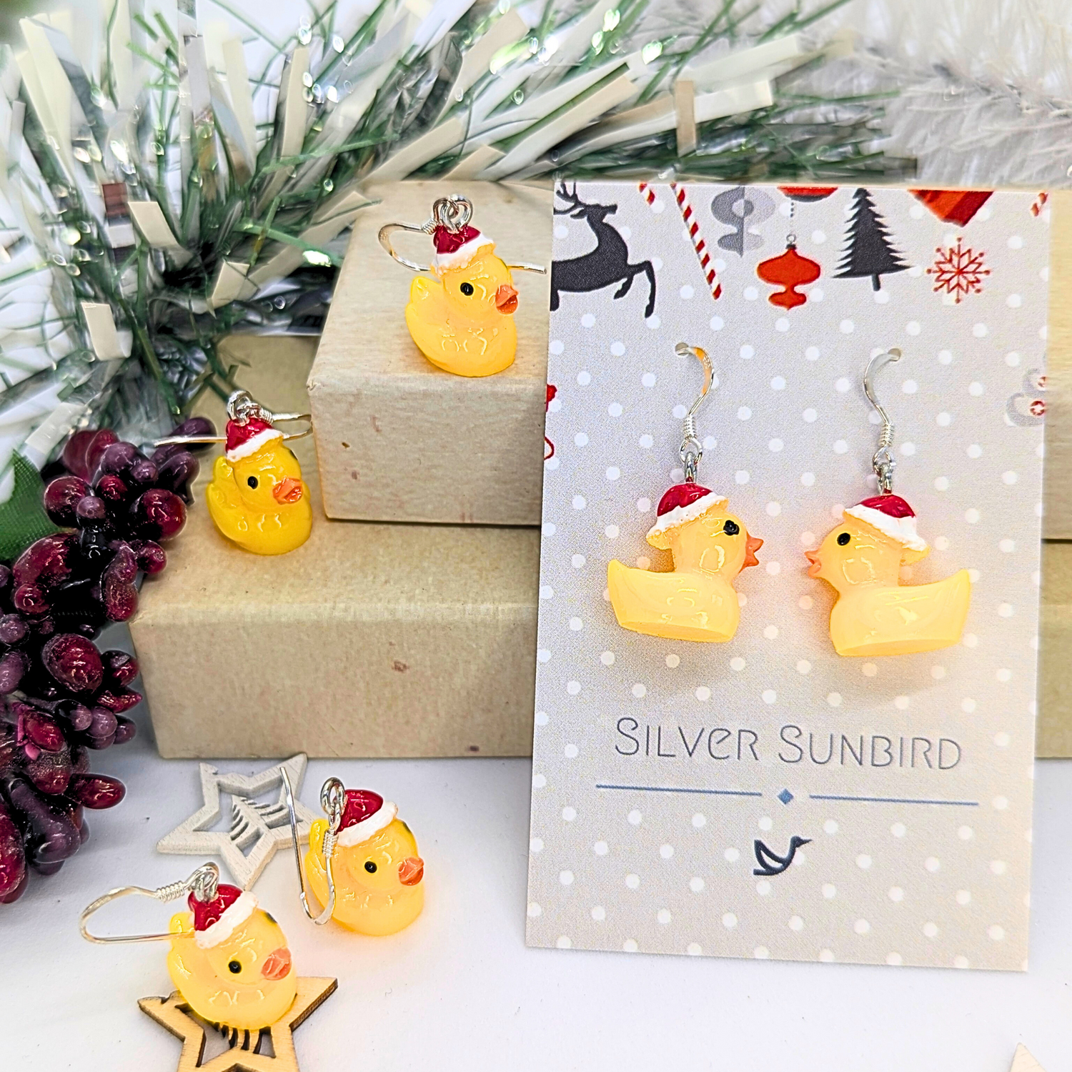 A close-up showcases the Silver Sunbird Festive Duck Earrings, featuring handcrafted yellow ducks adorned with Santa hats, elegantly hanging from sterling silver hooks. These earrings are presented on a cheerful card embellished with a pine branch and red berries in the background, making them an ideal holiday statement piece.