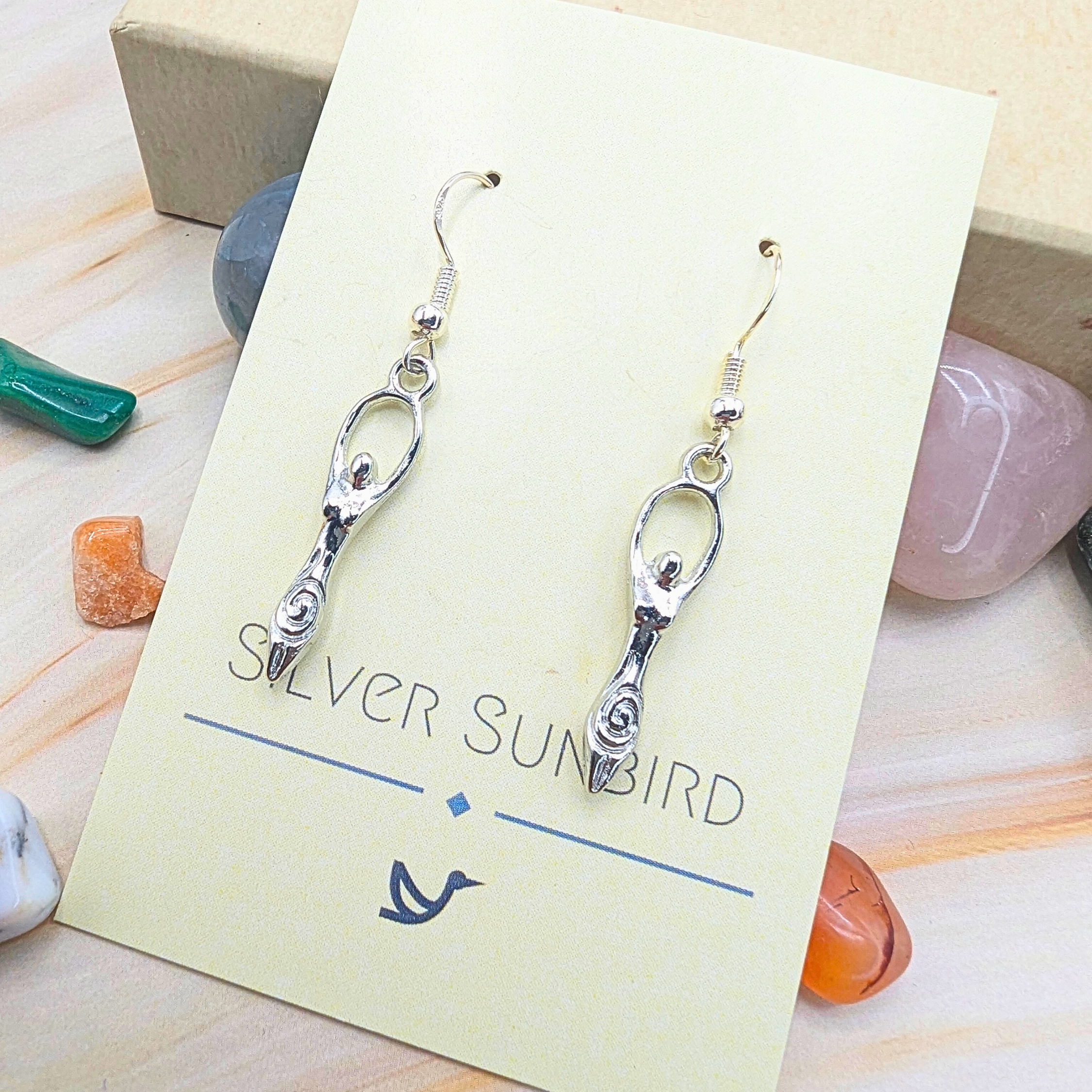 Fertility Goddess Dangle Earrings by Silver Sunbird are showcased on a cream card, surrounded by colorful polished stones atop a light wooden surface.