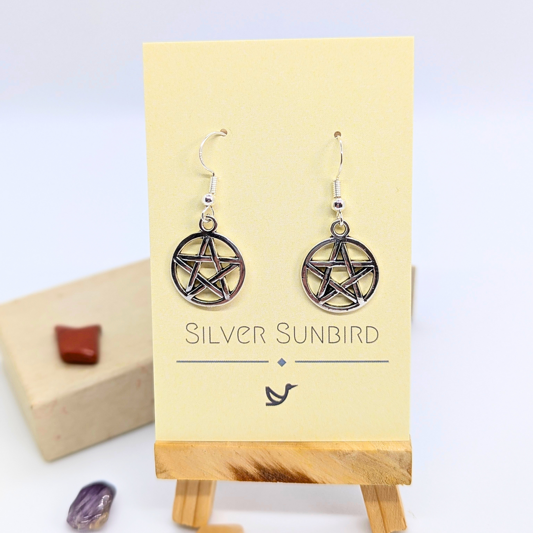 Pentagram Earrings by Silver Sunbird are shown on a branded card featuring a bird logo, with small gemstones and a wooden block in the background, emphasizing these Wicca jewelry pieces&