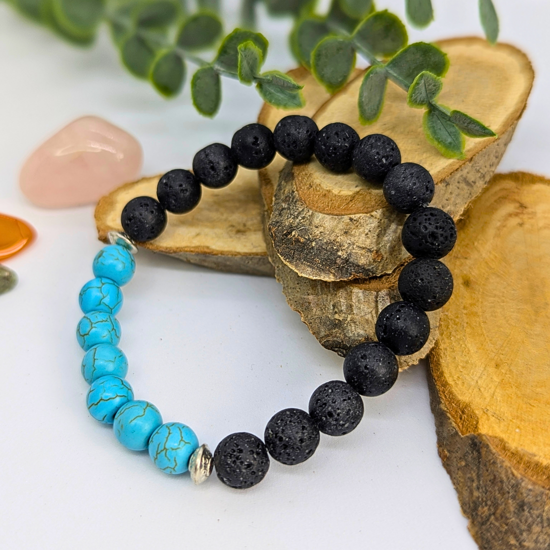 The Silver Sunbird Turquoise and Lava Stone Bracelet, featuring black lava stones and five turquoise beads, is displayed on wooden slices with blurred greenery and crystals in the background.