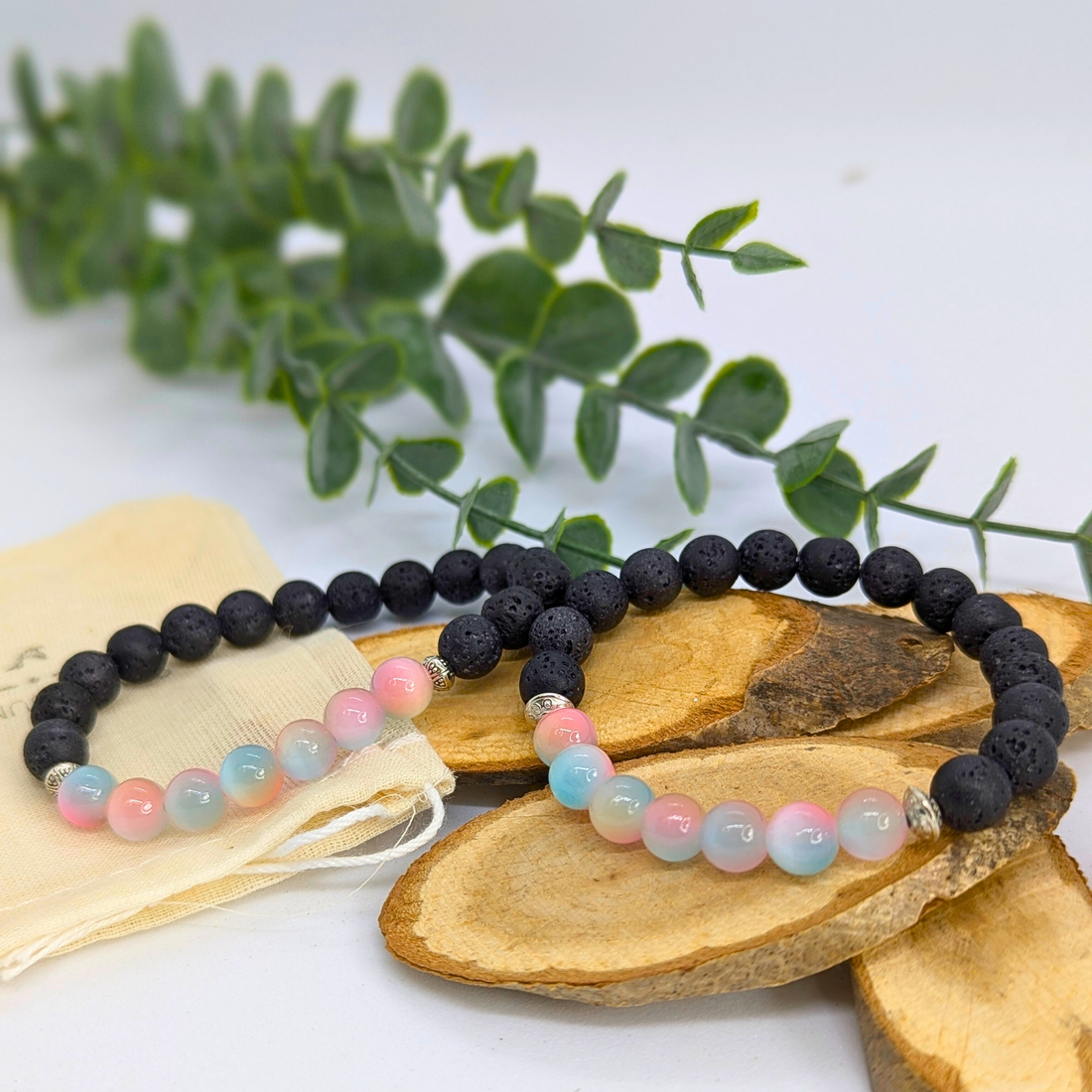 Two pastel Mermaid-Inspired Lava Stone Bracelets by Silver Sunbird, with black lava beads and blue-pink hues, rest on wooden slices. Perfect for ocean lovers, they evoke seaside charm and come with a fabric pouch, displayed beside a leafy branch.