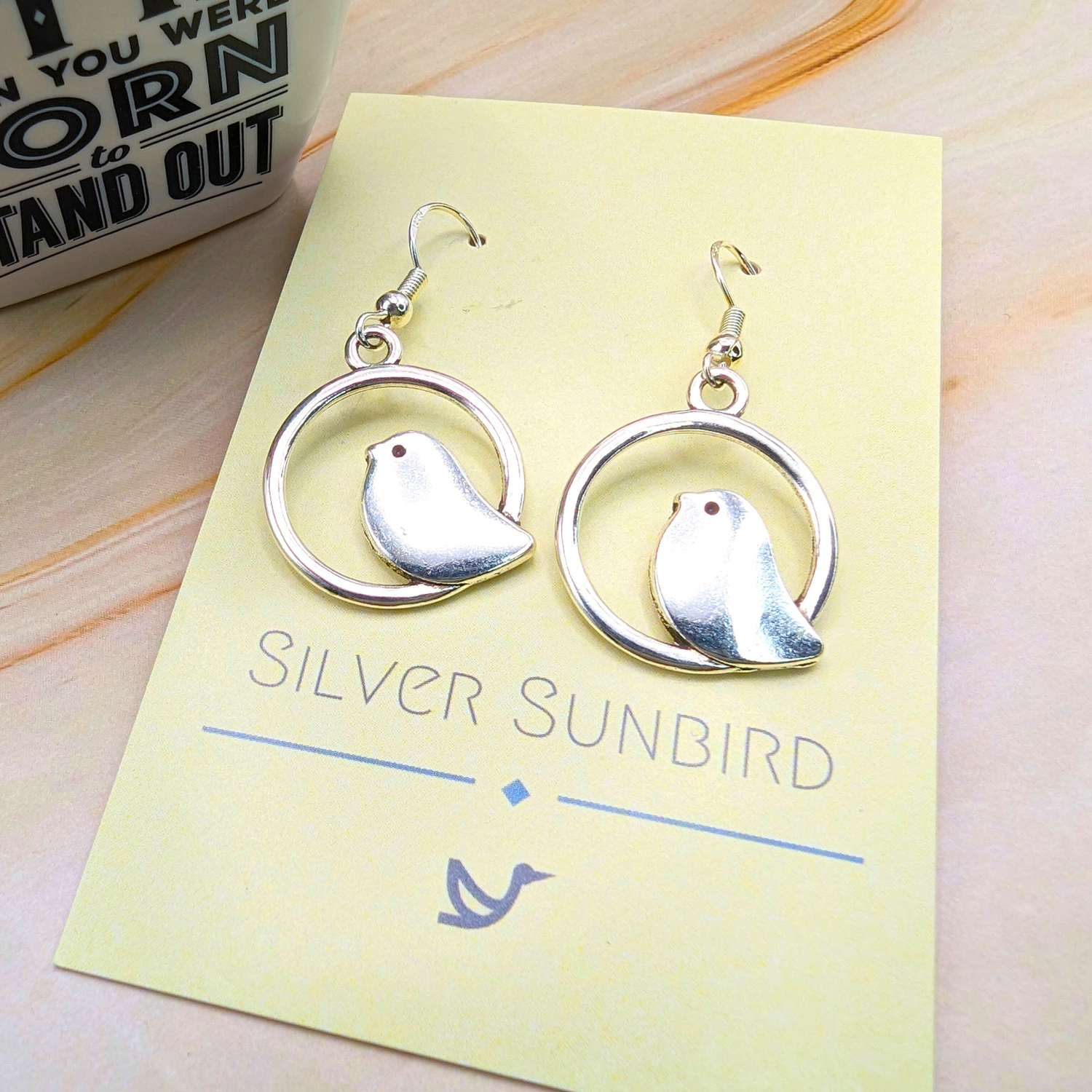 A pair of Beautiful Bird Earrings by Silver Sunbird, crafted from 925 Sterling Silver with a bird silhouette in a circular frame, is displayed on a "Silver Sunbird" card. Perfect for bird lovers, they rest on a light surface beside an inspirational mug.