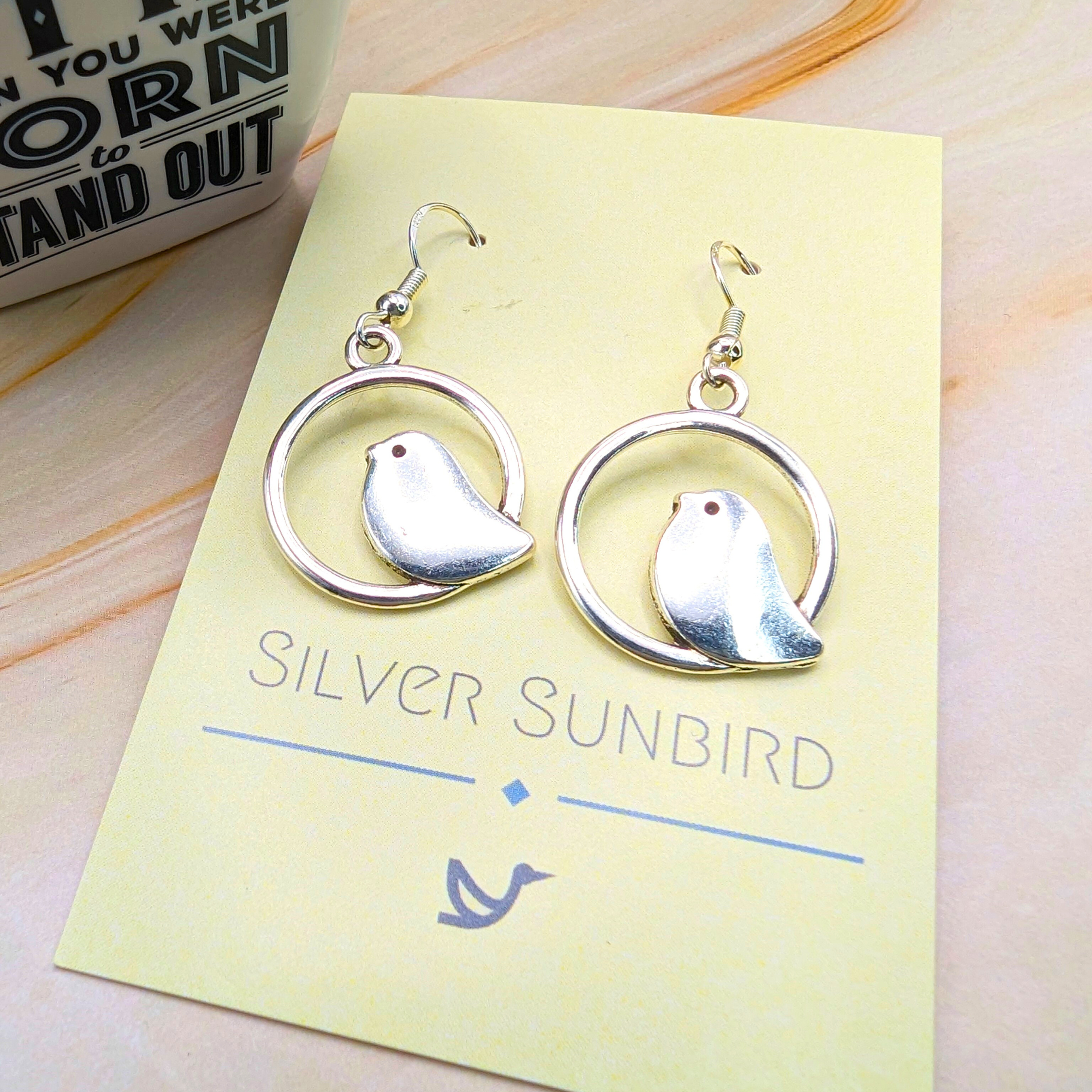 A pair of Beautiful Bird Earrings by Silver Sunbird, crafted from 925 Sterling Silver with a bird silhouette in a circular frame, is displayed on a "Silver Sunbird" card. Perfect for bird lovers, they rest on a light surface beside an inspirational mug.