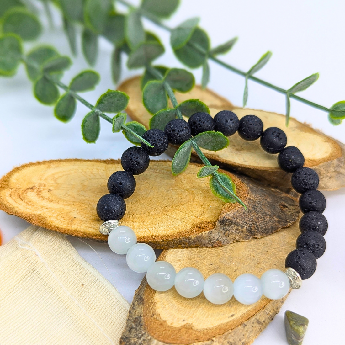 The Silver Sunbird Moonstone and Lava Bracelet, featuring black lava stone and light blue moonstone beads, is shown on wooden slices with green sprigs and a drawstring pouch nearby.