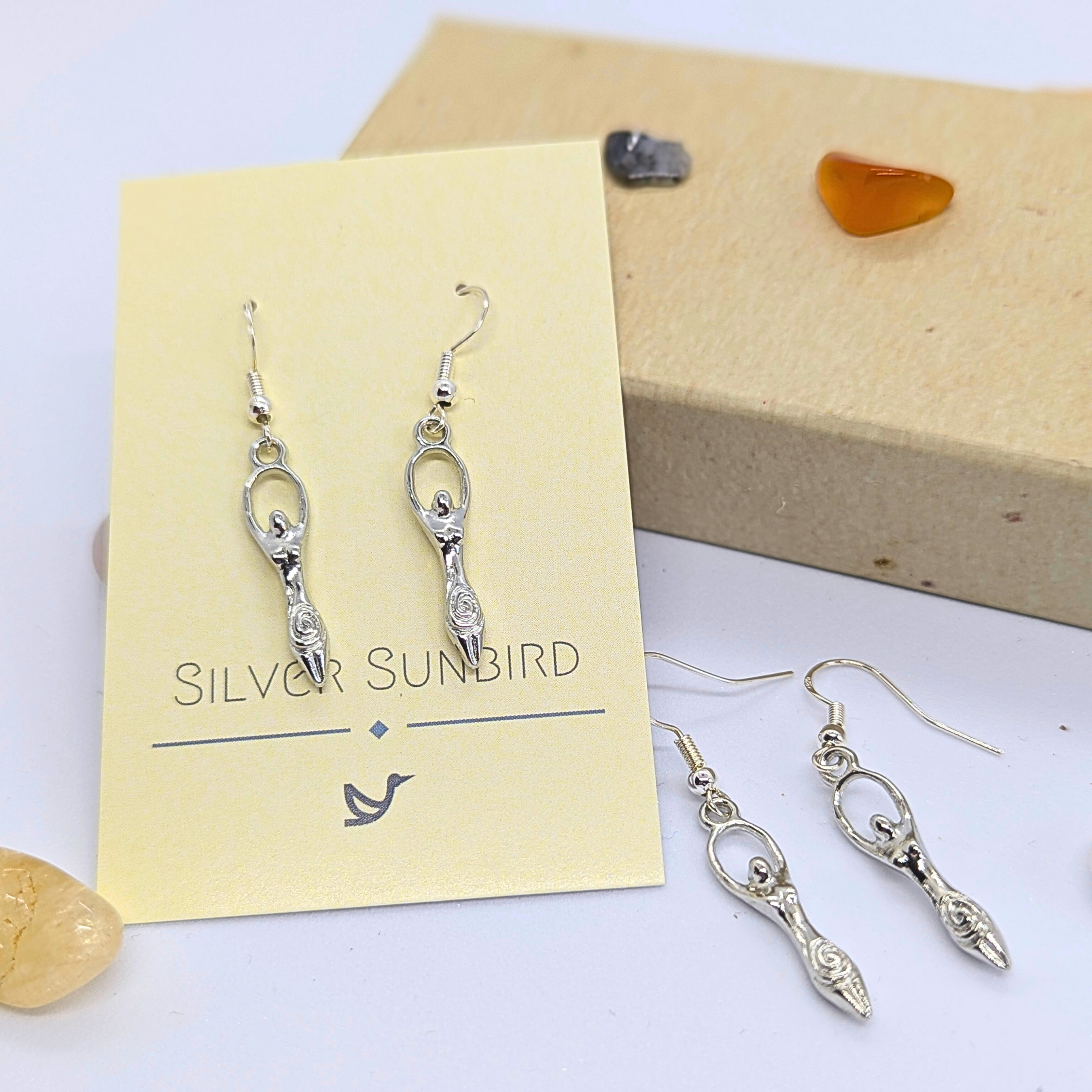 A pair of Silver Sunbird Fertility Goddess Dangle Earrings is shown on a cream card, with another matching pair nearby amid small tumbled stones and a brown box.