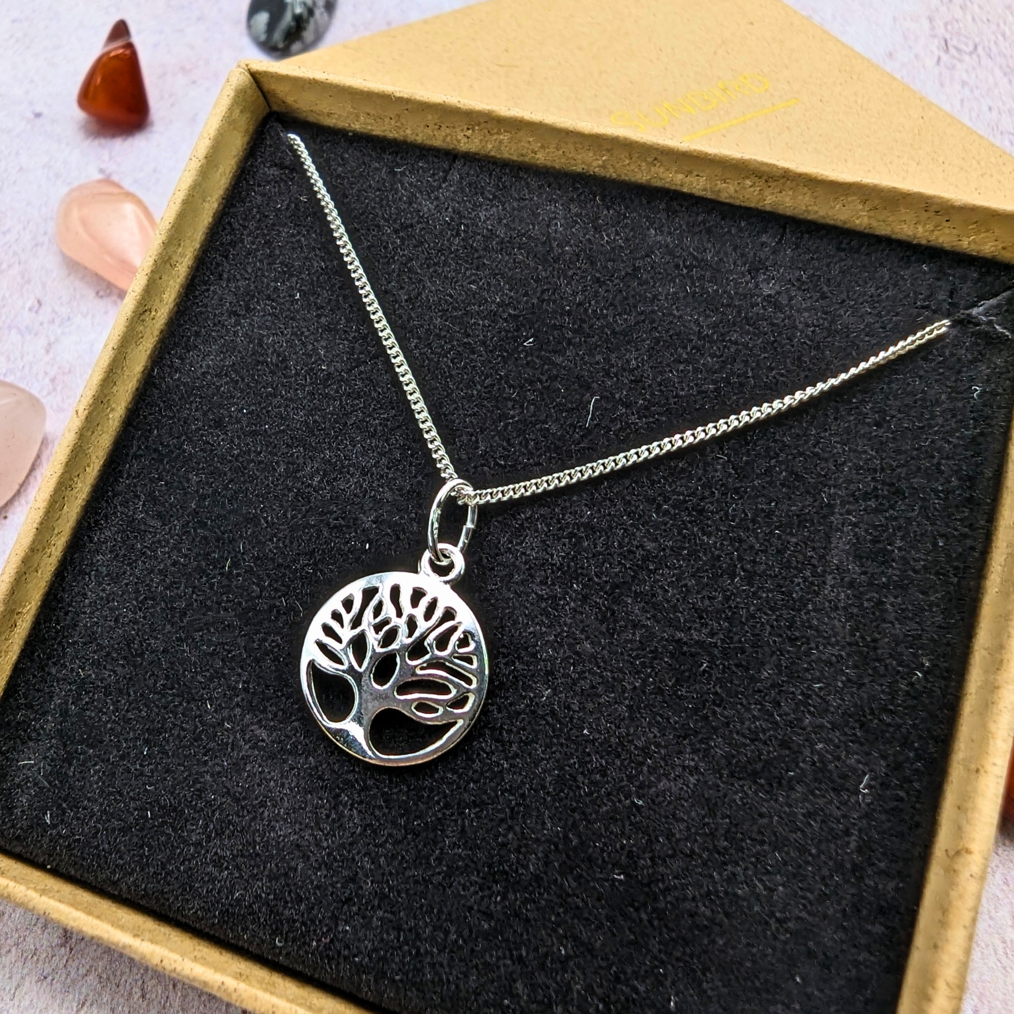 The Silver Sunbird Tree of Life Sterling Silver Necklace, featuring a round pendant, rests in a black velvet-lined box. Colorful polished gemstones nearby highlight this meaningful jewelry piece.