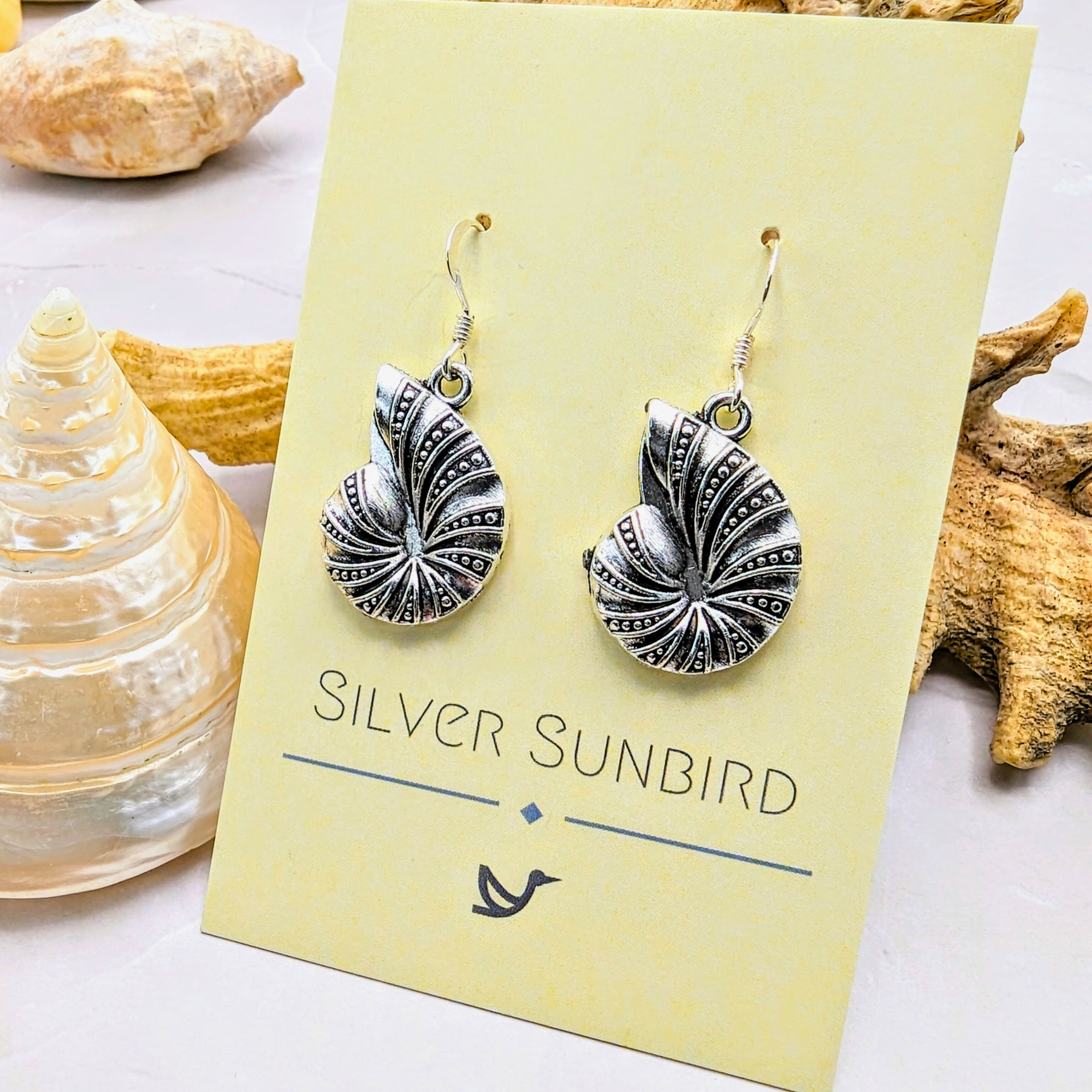 The Shoreline Spiral Shell Earrings by Silver Sunbird, featuring intricate silver patterns, are displayed on a yellow brand card amid seashells and coral, highlighting this ocean-inspired jewellery.