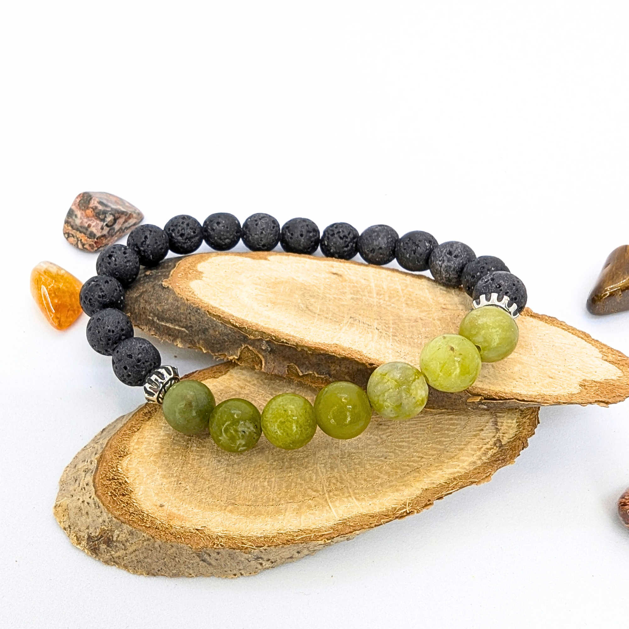 The Silver Sunbird Peridot and Lava Stone Bracelet features alternating black and green beads, shown on stacked wooden slices with vibrant polished stones against a white background.