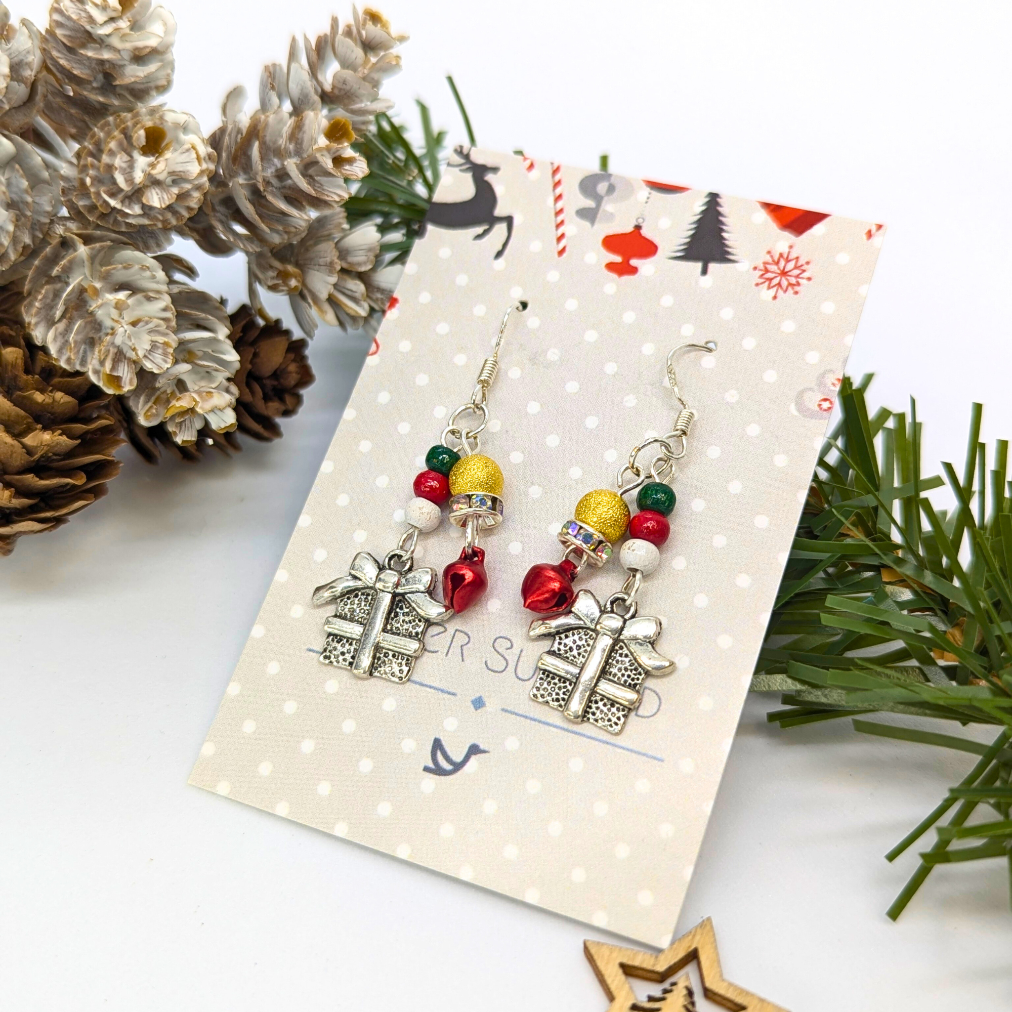 Silver Sunbird’s Gift Box Dangle Earrings feature colorful beads on sterling silver hooks, presented on a Christmas card and surrounded by festive pinecones and greenery for a cheerful seasonal display.