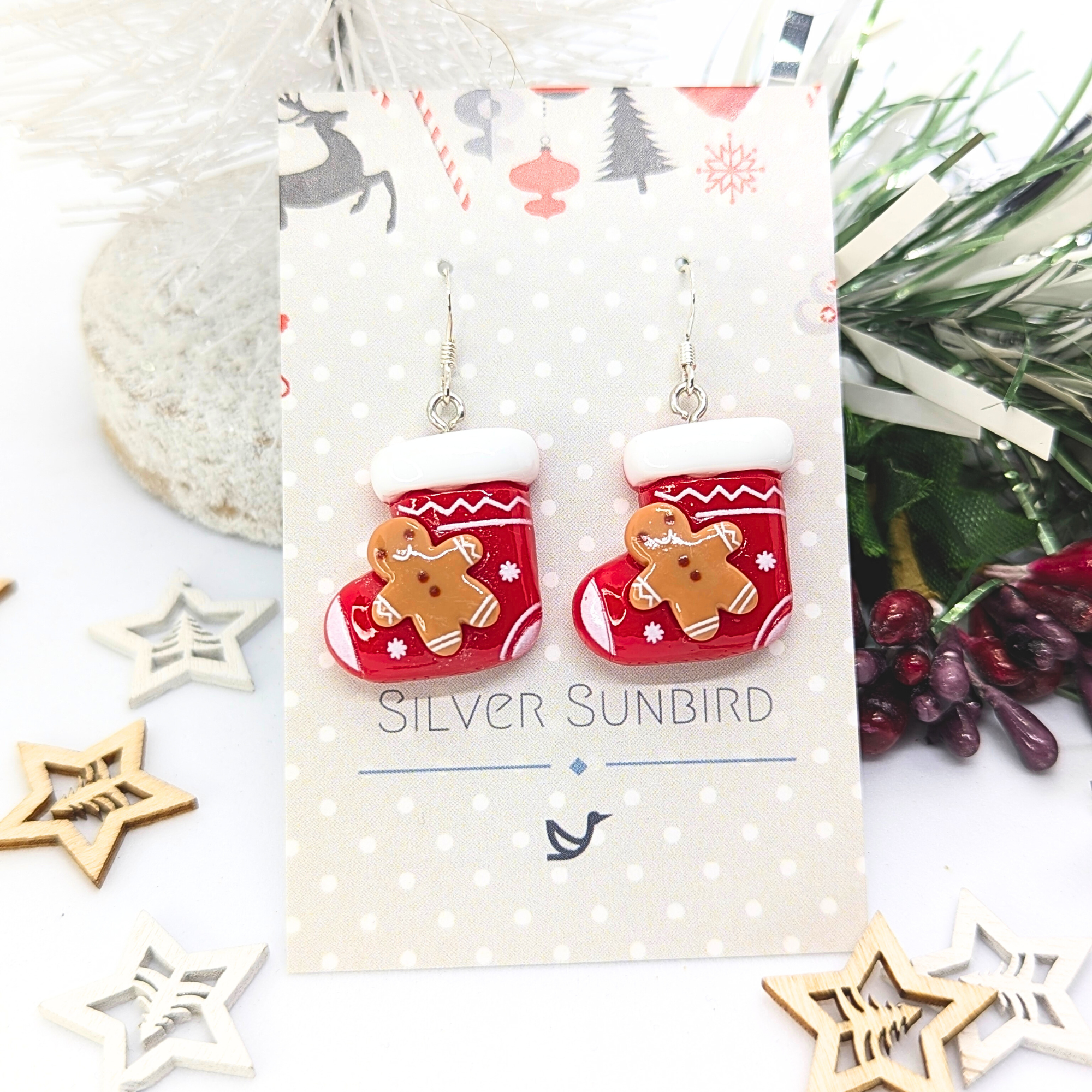 Displayed on an elegant "Silver Sunbird" decorative card, the Gingerbread Men Stocking Earrings feature a festive design with white trim and gingerbread figures. Surrounded by holiday-themed decorations and foliage, they are the perfect holiday accessory.
