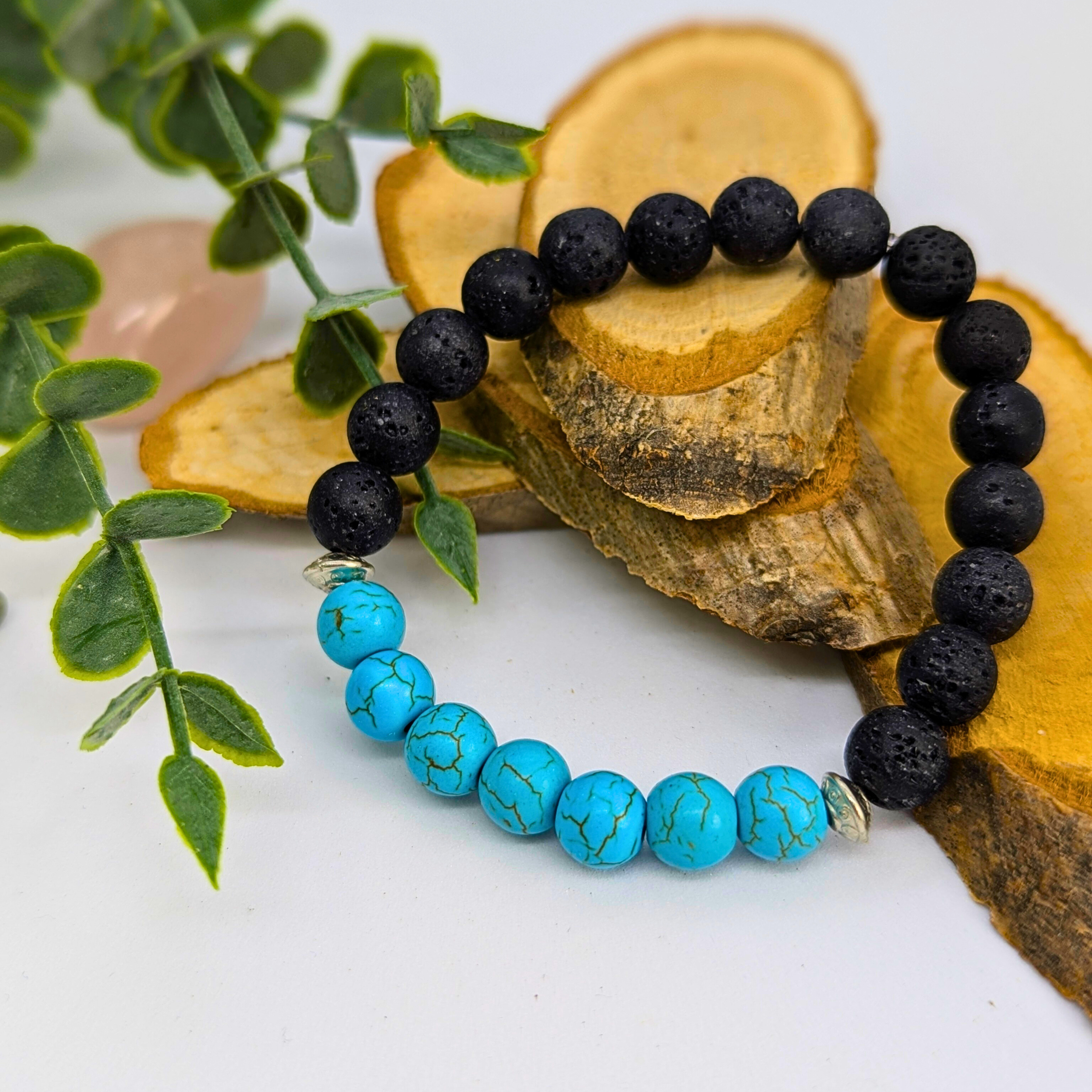 The Silver Sunbird Turquoise and Lava Stone Bracelets are displayed on layered wooden pieces beside green leaves, set against a white background.