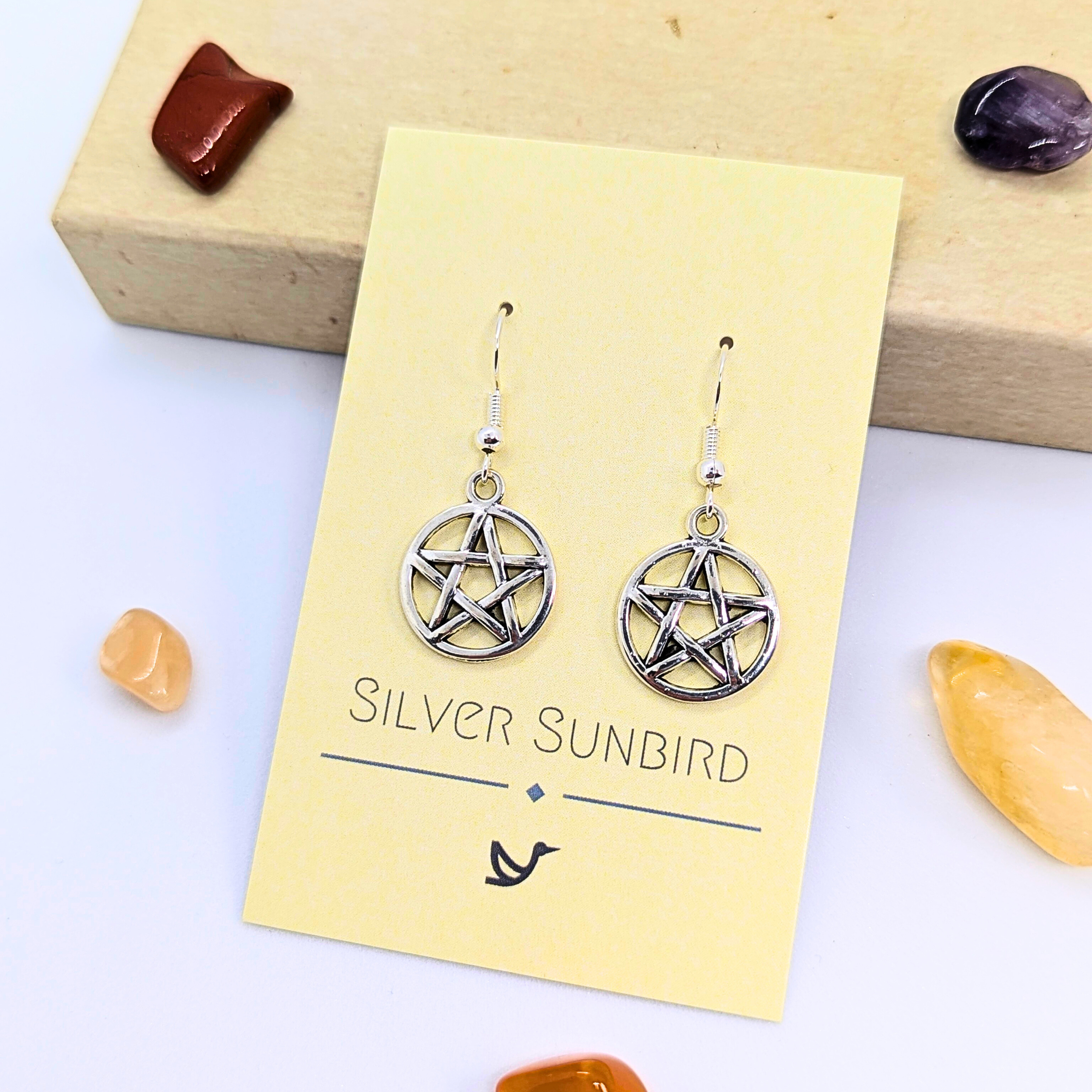 Pentagram Earrings by Silver Sunbird are displayed on a branded card, surrounded by polished gemstones. These striking Wicca jewelry pieces celebrate pagan symbolism on a white surface.