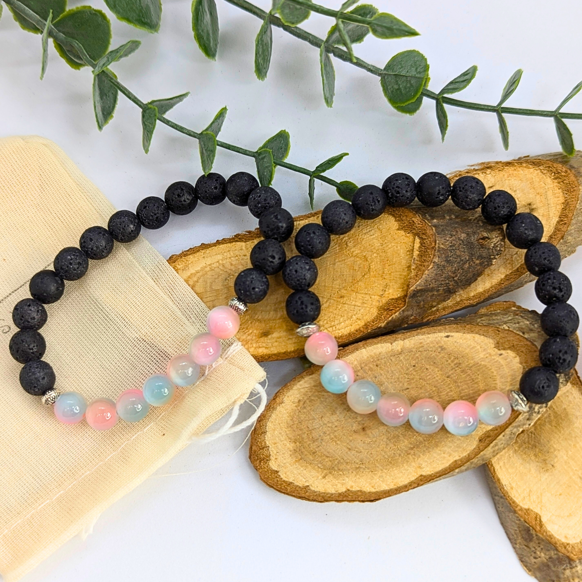 The Silver Sunbird Mermaid-Inspired Lava Stone Bracelet, featuring pastel beads with black lava stones and pink and blue accents, rests on wooden slices. A cloth pouch and green leafy branches complete the serene setting.