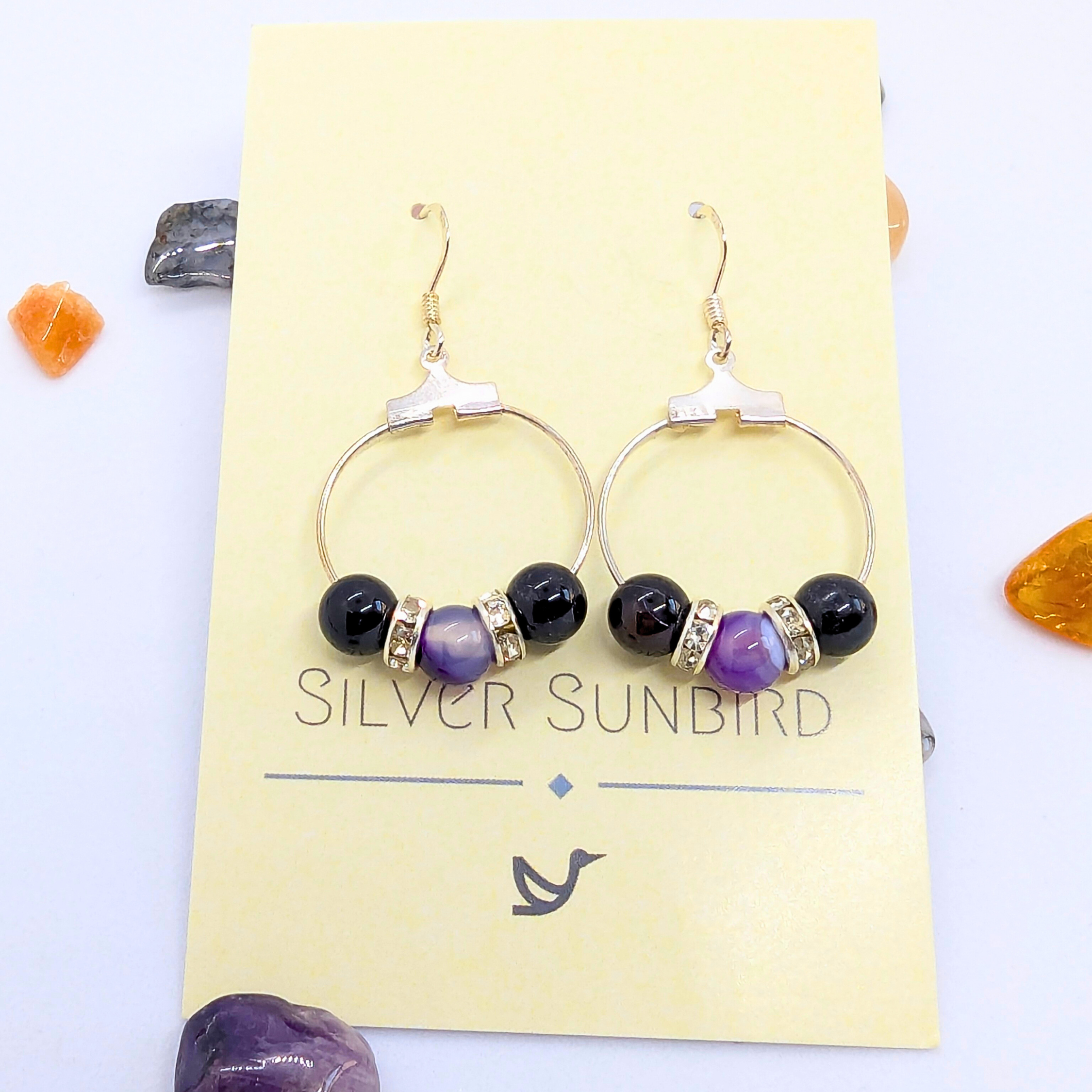 A pair of elegant Amethyst Large Statement Earrings displayed on a yellow card labelled "Silver Sunbird." The earrings feature purple amethyst round gemstones, accented with silver rhinestone spacers. The card is adorned with small decorative stones in various colors around its edges.