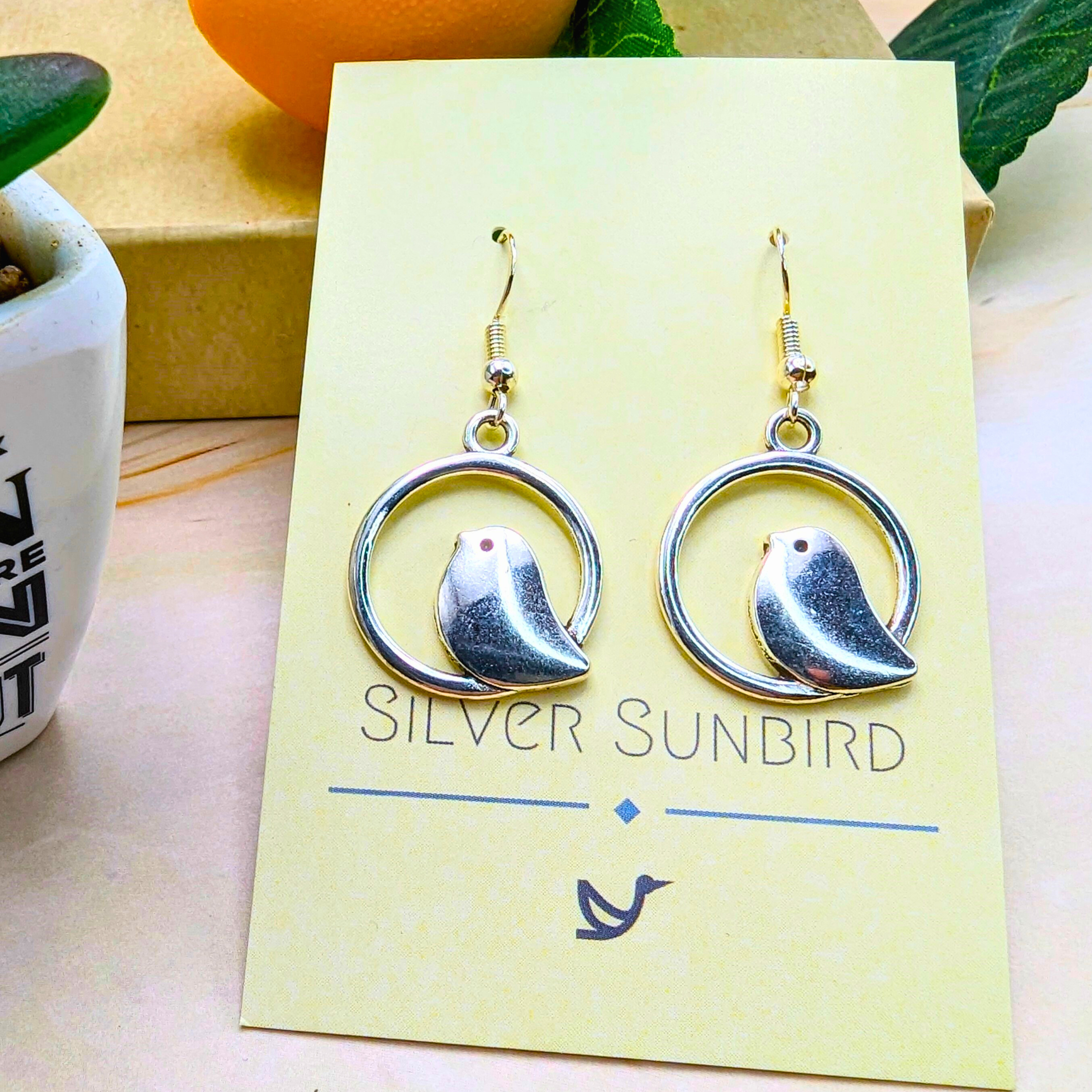 Beautiful Bird Earrings from Silver Sunbird, made of 925 Sterling Silver, feature bird shapes within circular frames. Displayed on a card labeled "Silver Sunbird" and set on a wooden surface, these earrings are a delightful choice for bird lovers with an eye for elegant accessories.