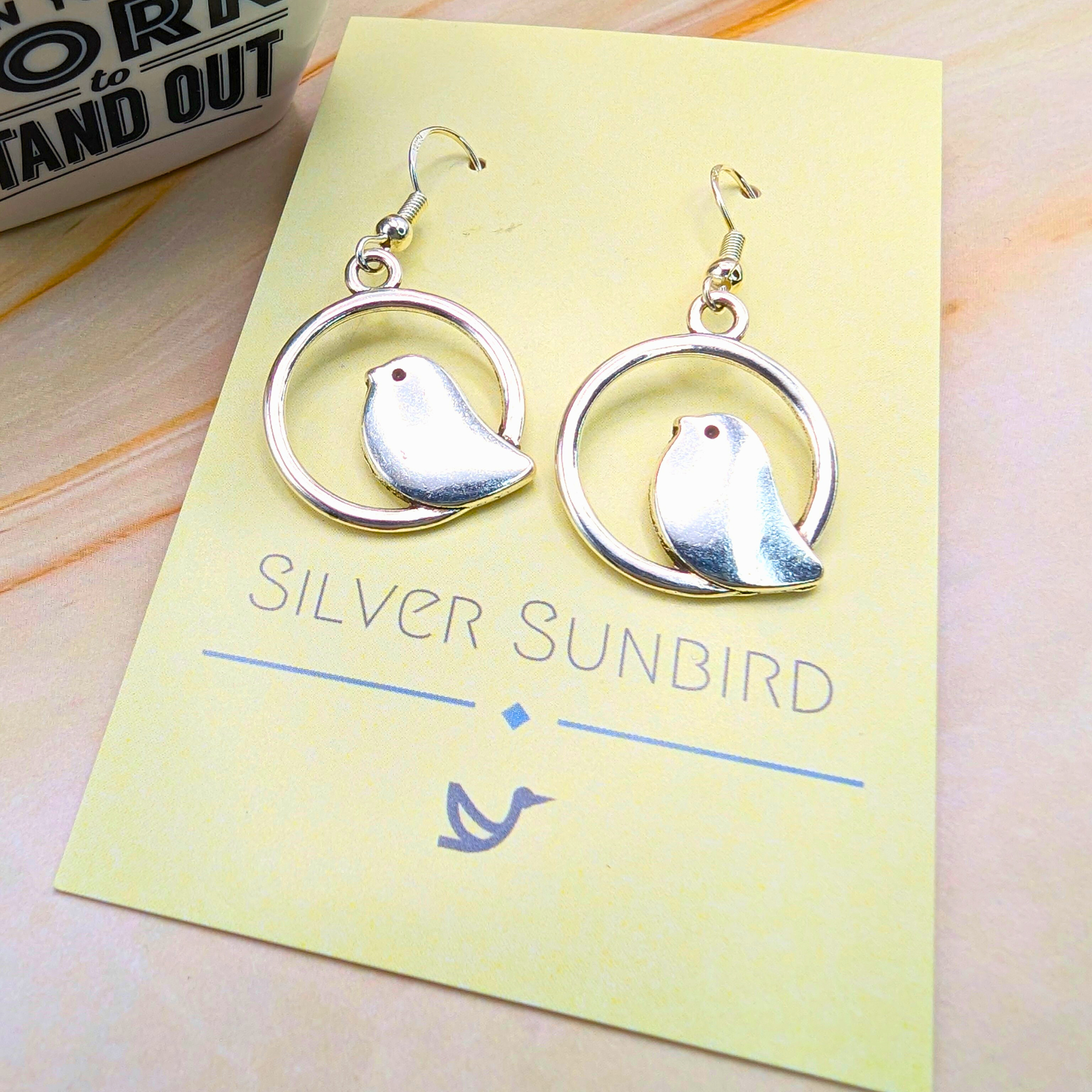 A pair of Beautiful Bird Earrings by Silver Sunbird, crafted from 925 Sterling Silver, sits on a yellow card with "Silver Sunbird" branding and bird logo. Ideal for bird lovers, these earrings showcase delicate bird shapes within circular frames.