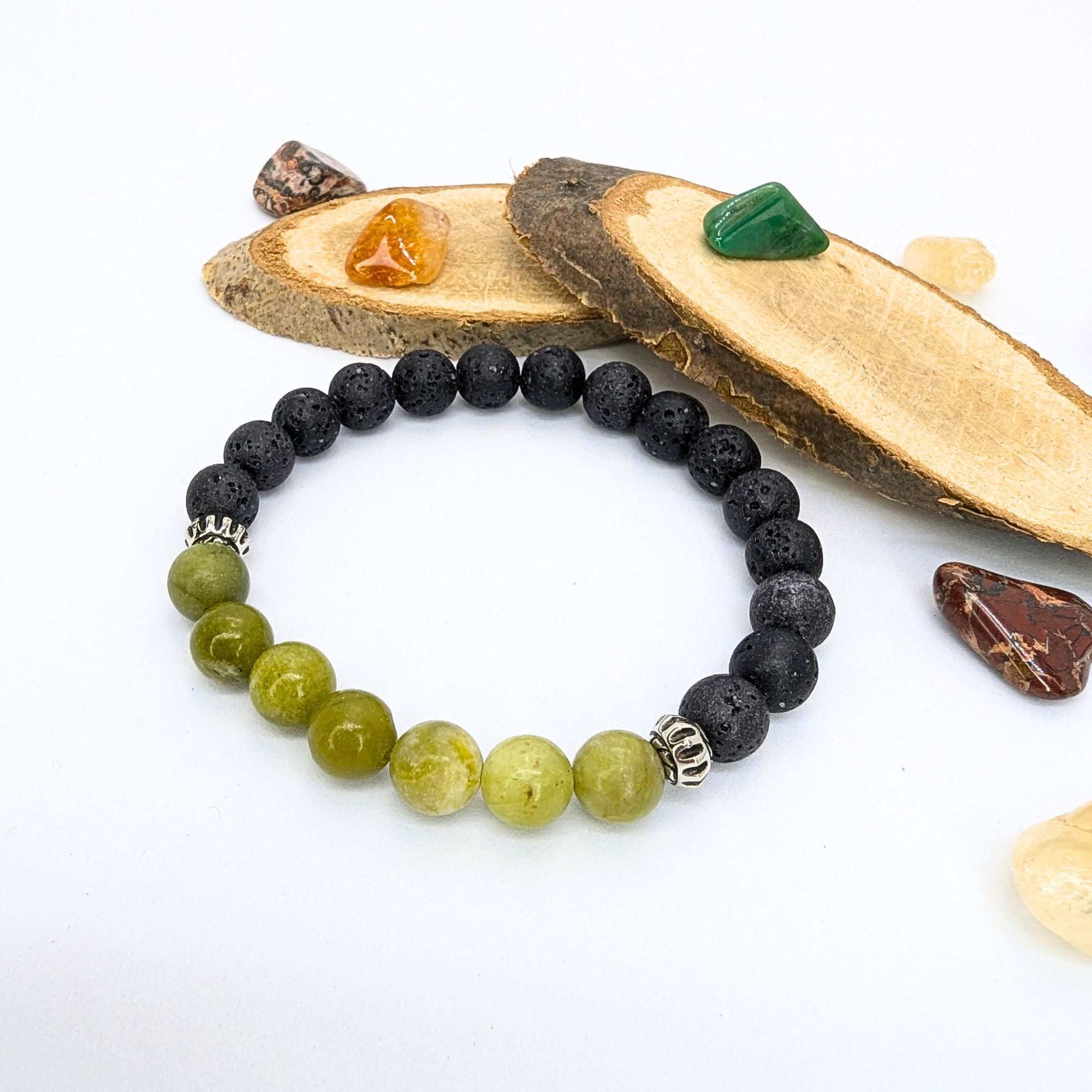 The Silver Sunbird Peridot and Lava Stone Bracelet, featuring alternating black and green beads with two silver spacers, is showcased on a white surface with wooden slices and colorful polished stones in the background.