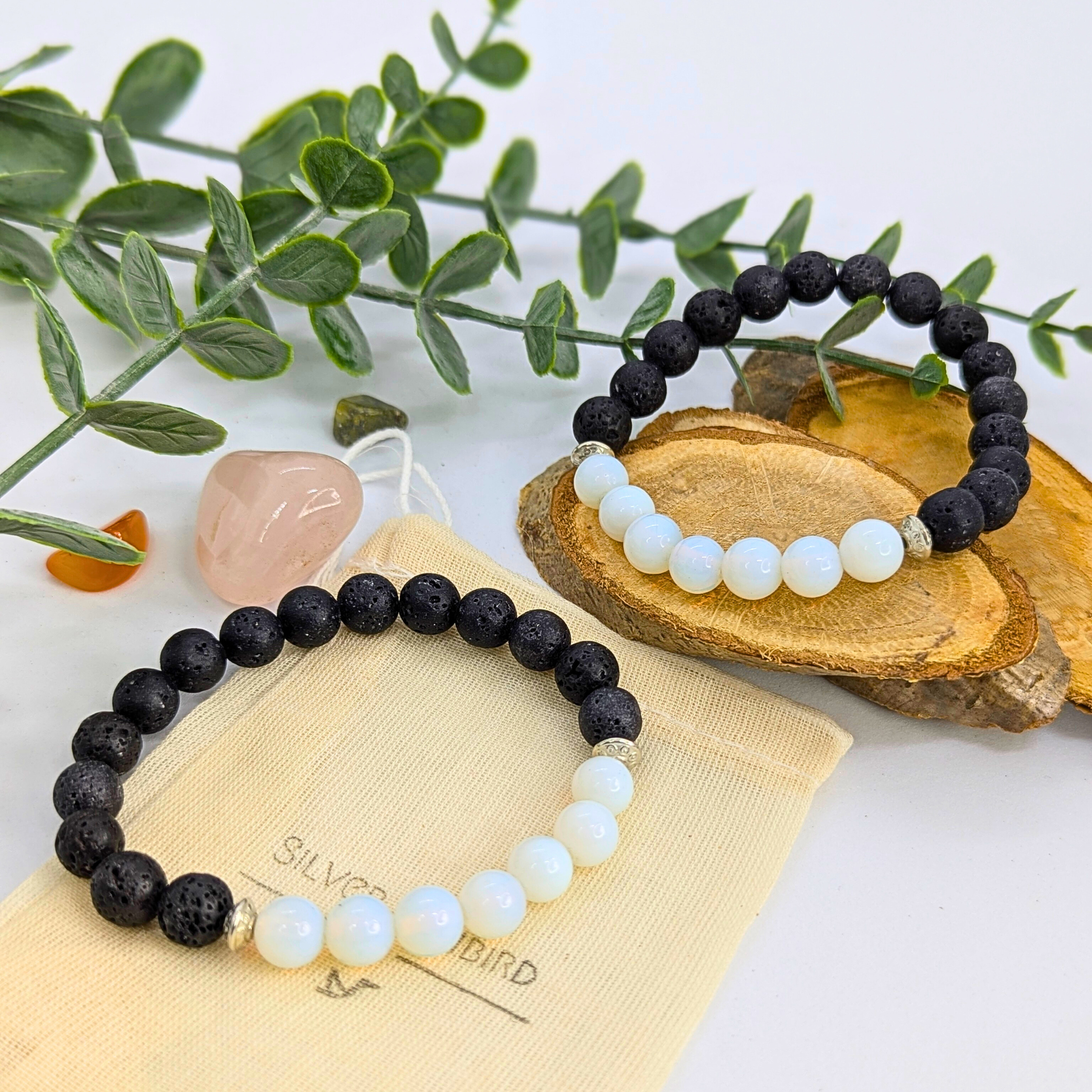 Two Silver Sunbird Opal and Lava Stone Bracelets rest on a wooden slab and pouch, with greenery and two crystals—one pink, one orange—in the bright, minimal background.