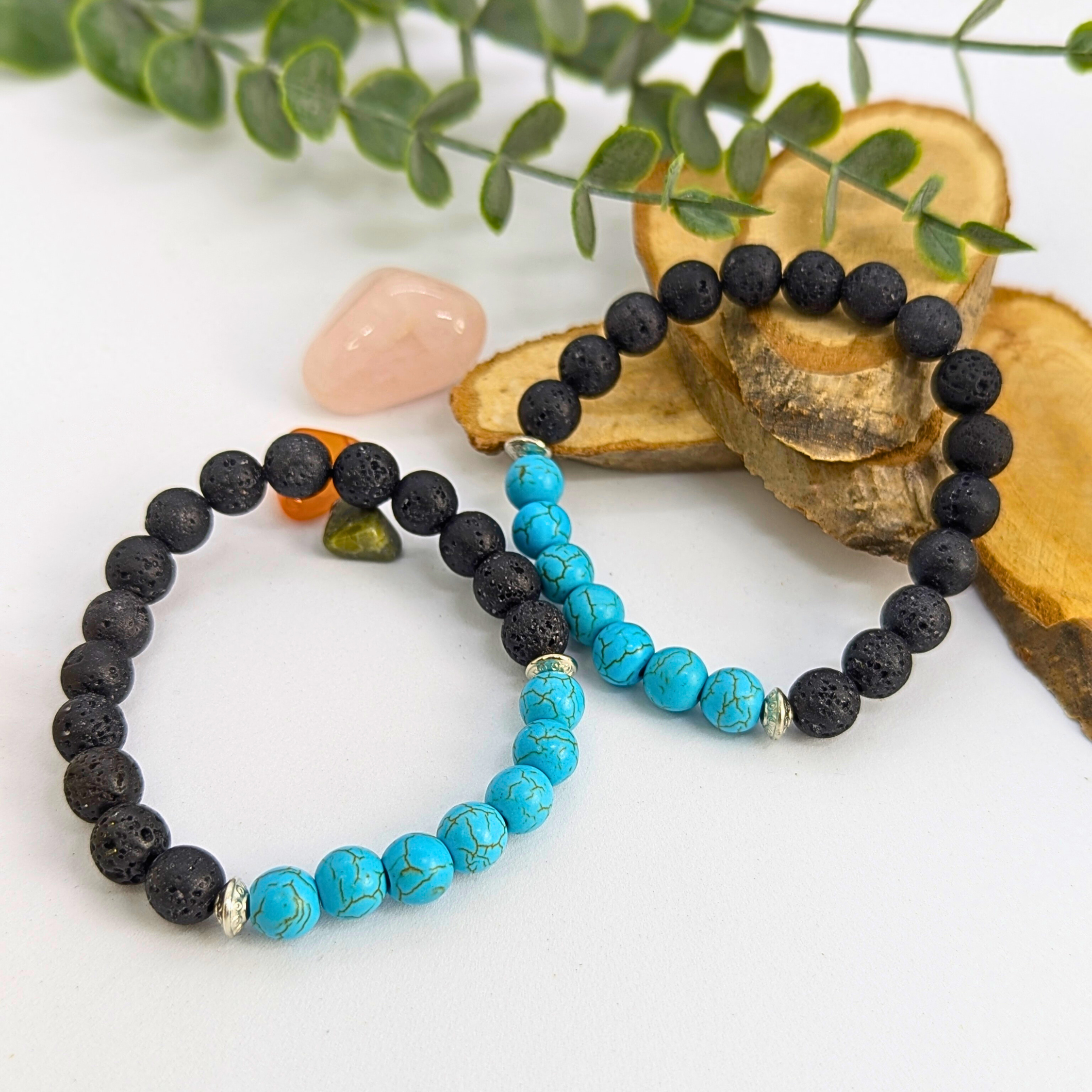 Two Turquoise and Lava Stone Bracelets by Silver Sunbird are displayed on sliced wood rounds with green foliage. One features black and turquoise beads, the other has black beads with a few small colored stones nearby.