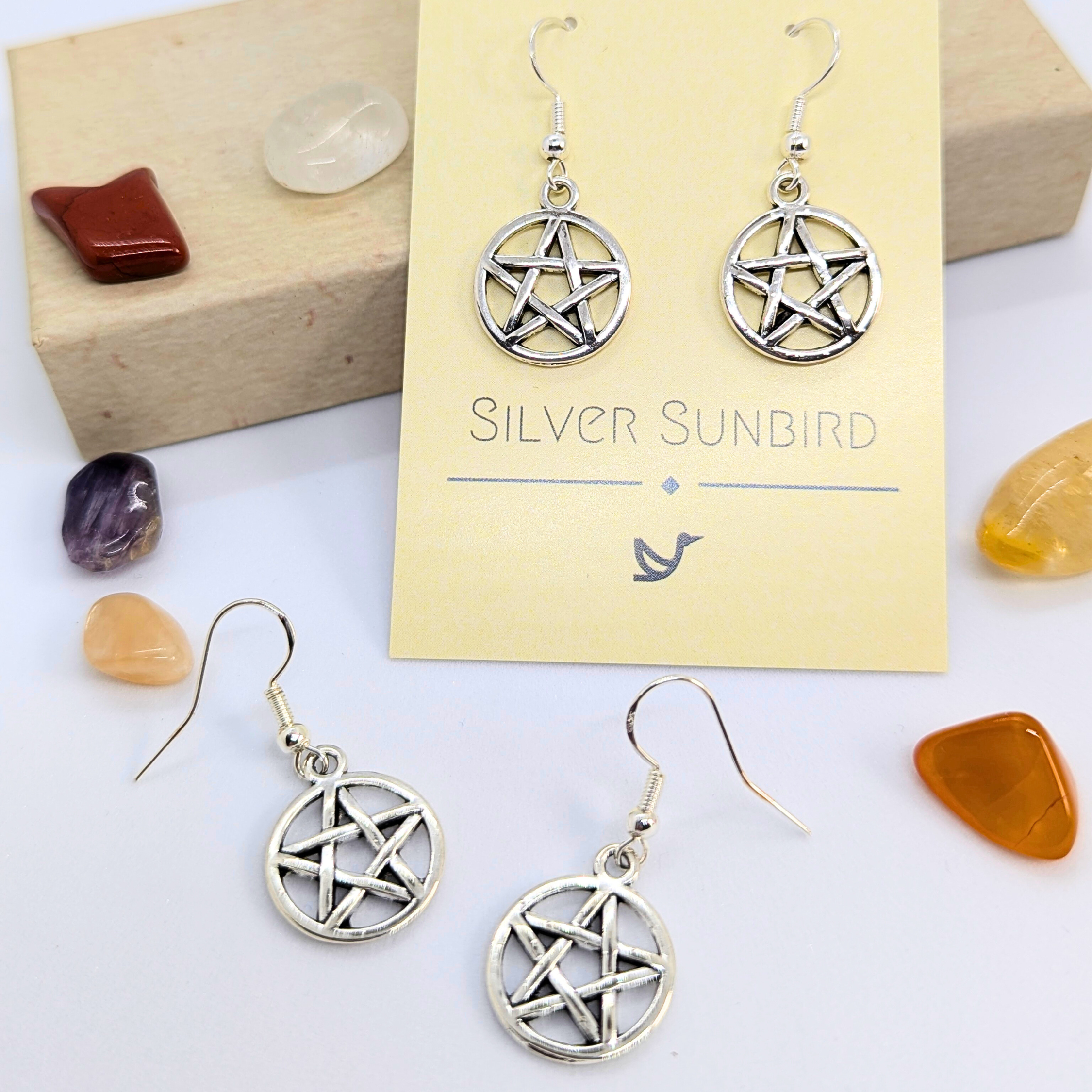 Two pairs of Silver Sunbird Pentagram Earrings, a bold Wicca jewelry piece, are shown with the brand card and surrounded by colorful polished stones on a white surface.